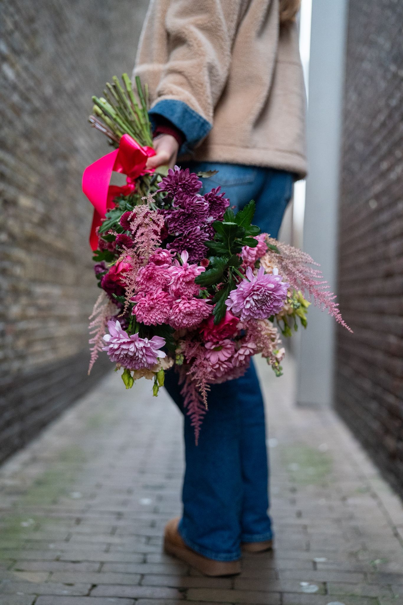 In een smal bakstenen steegje staat iemand in een beige jasje en spijkerbroek met een groot, opvallend roze en paars boeket bloemen.