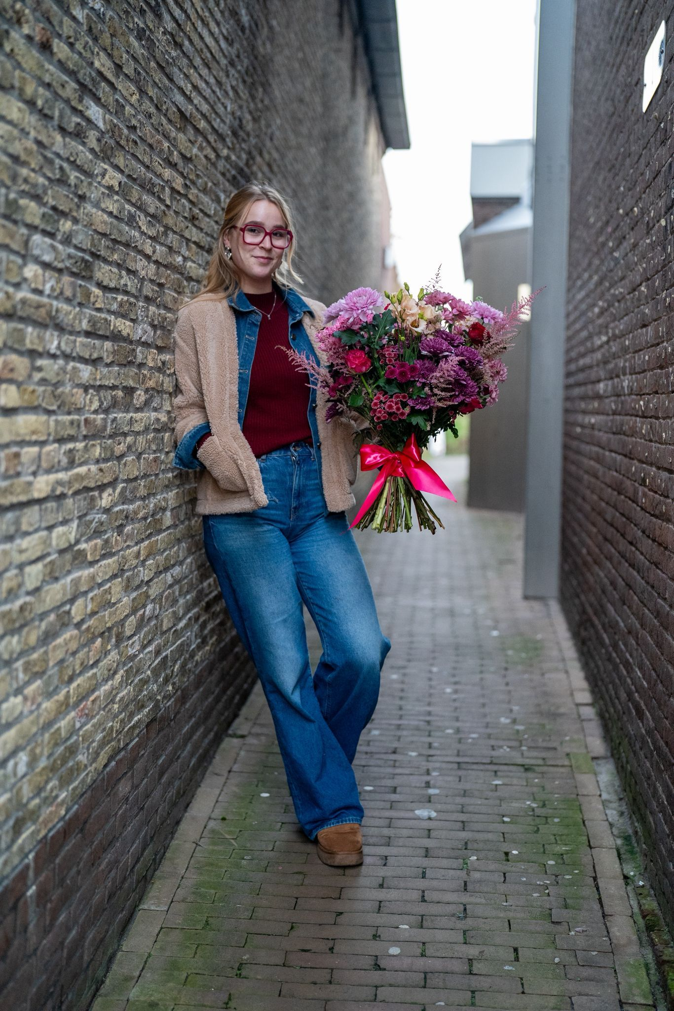 Een persoon die tegen een bakstenen muur in een steegje leunt, een groot boeket paarse bloemen vasthoudt en glimlacht.