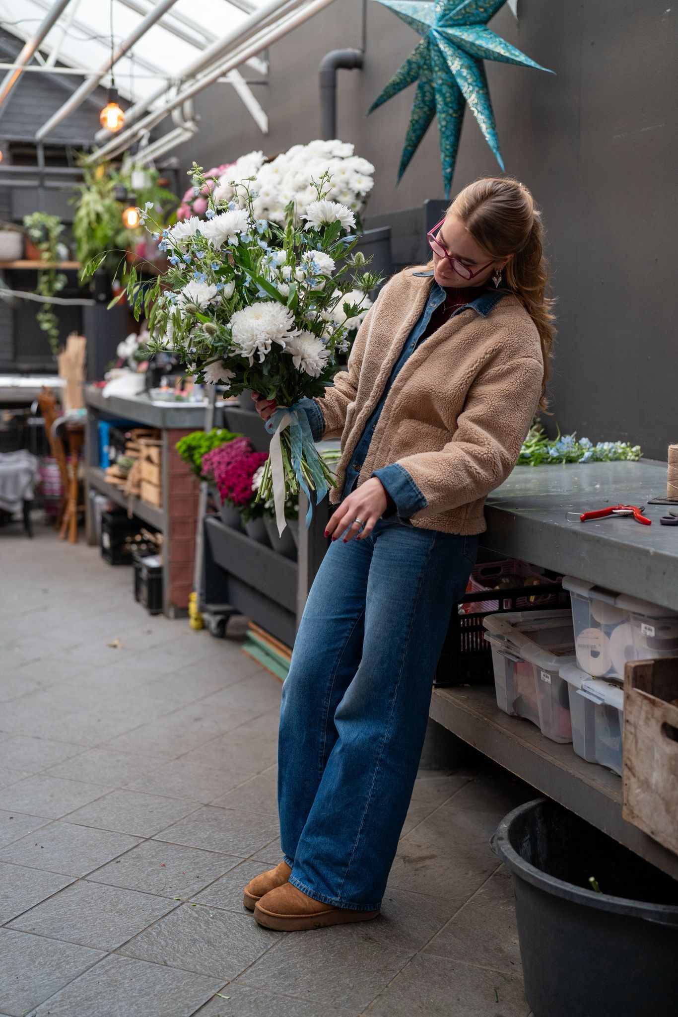 Een persoon in een beige, pluizige jas en een blauwe spijkerbroek leunt tegen een stenen aanrecht in een kas, naast een bos bloemen.