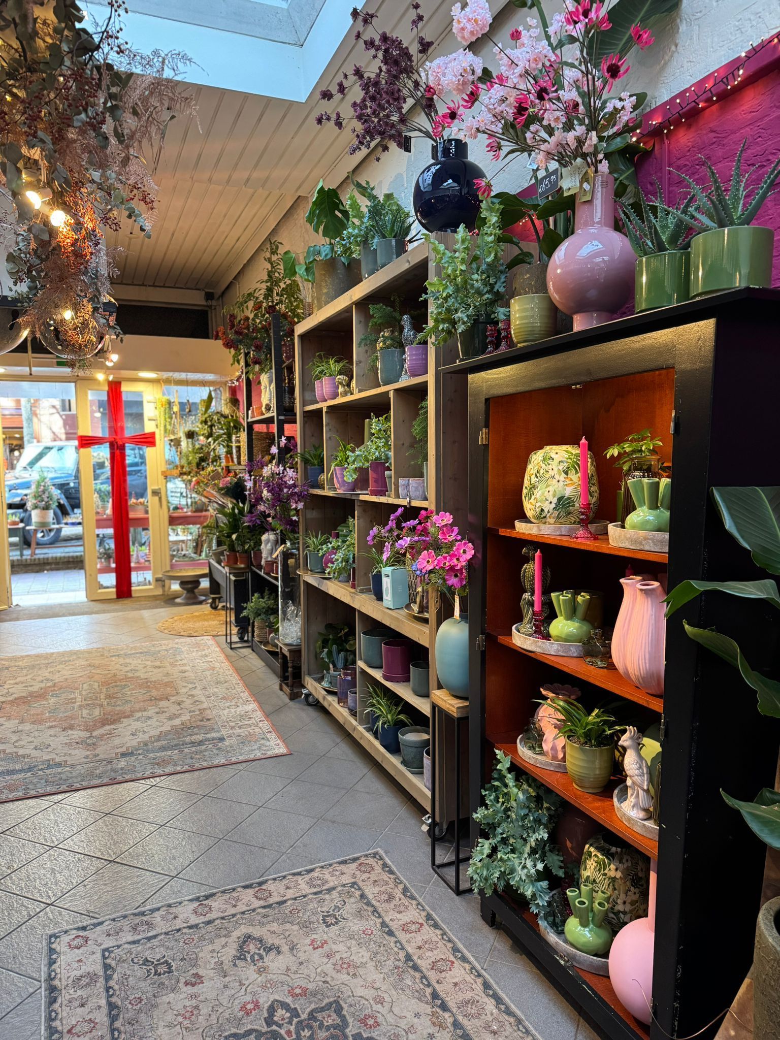 Een helder verlichte winkelruimte met houten schappen gevuld met diverse kamerplanten, bloemen en aardewerk.