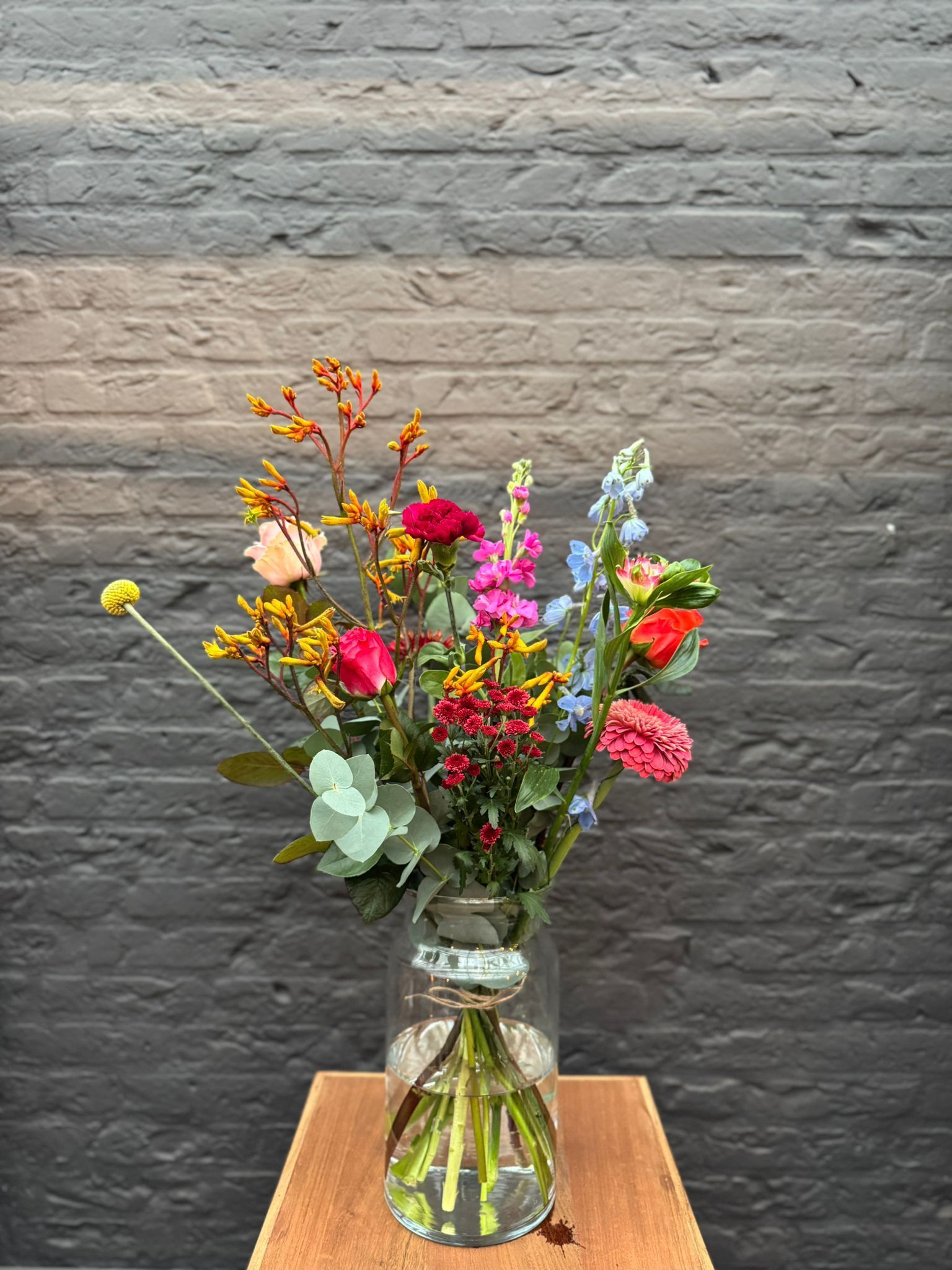 Een levendig boeket kleurrijke bloemen, waaronder rozen, lelies en blauwe riddersporen, in een transparante glazen vaas op een houten standaard.