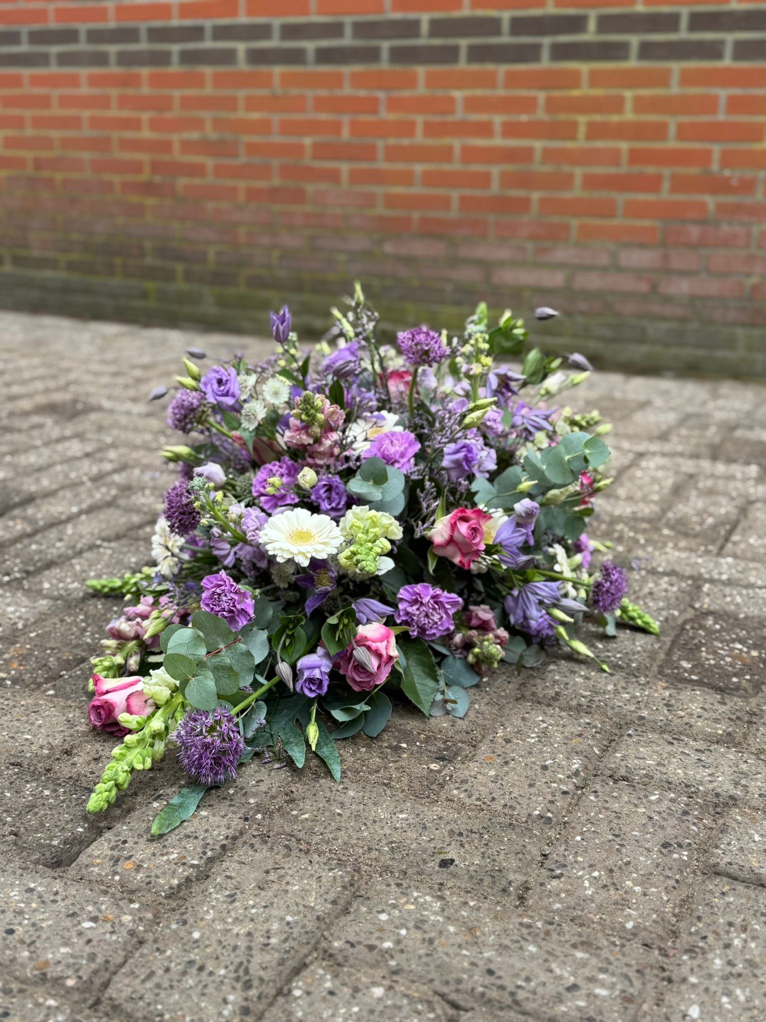 Een traanvormig bloemstuk met paarse, roze en witte bloemen en groen, geplaatst op een stenen terras.