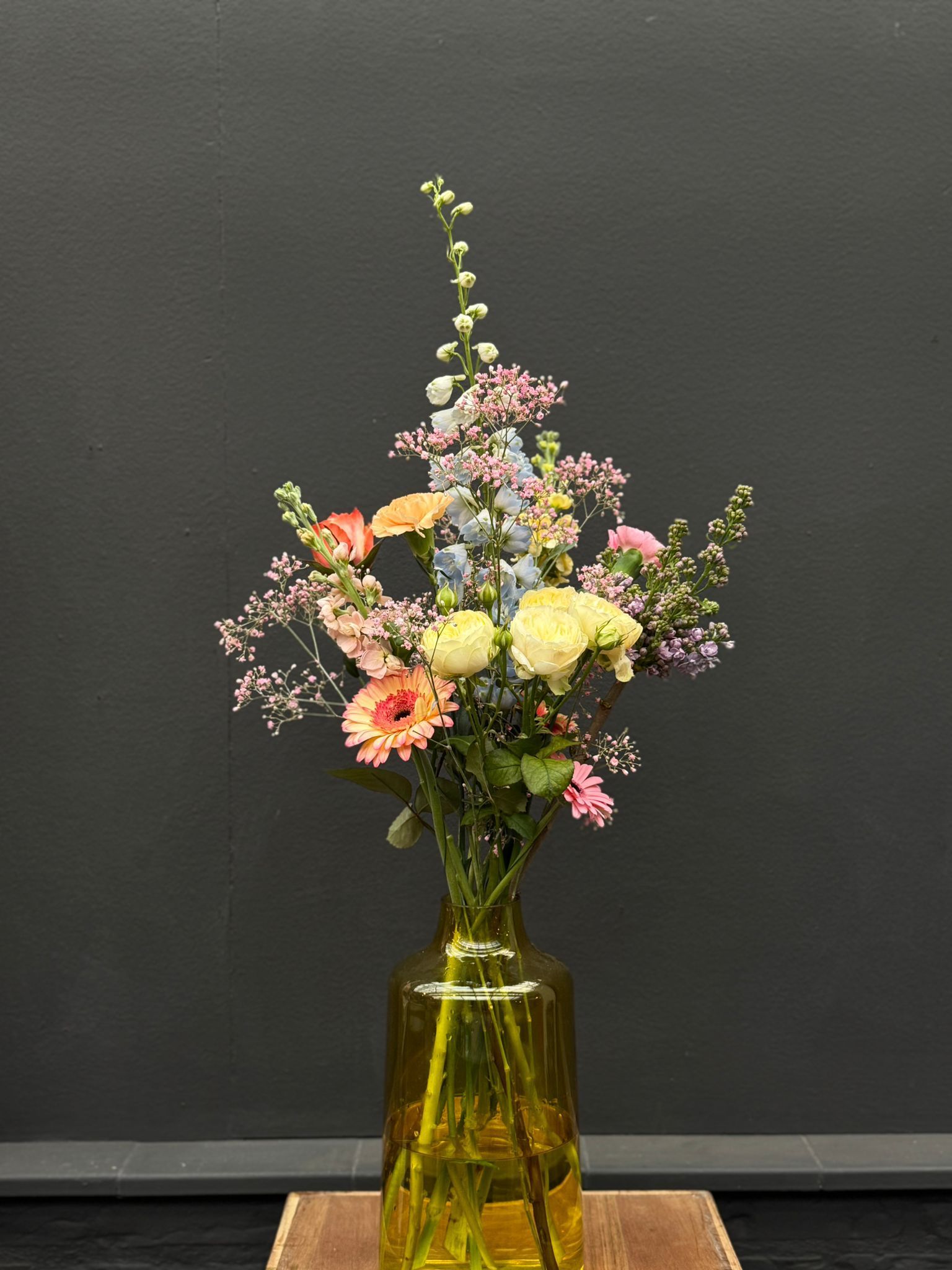 Een kleurrijk boeket pastelkleurige bloemen in een hoge, doorschijnende gele glazen vaas tegen een donkergrijze achtergrond.