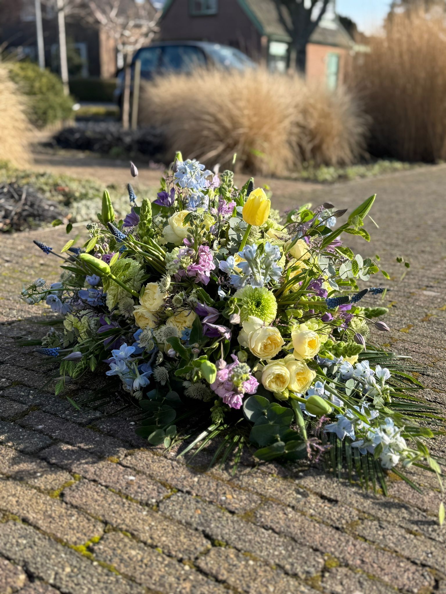 Een bloemstuk met gele, paarse en blauwe bloemen, geplaatst op een bestrate ondergrond in een buitenomgeving.