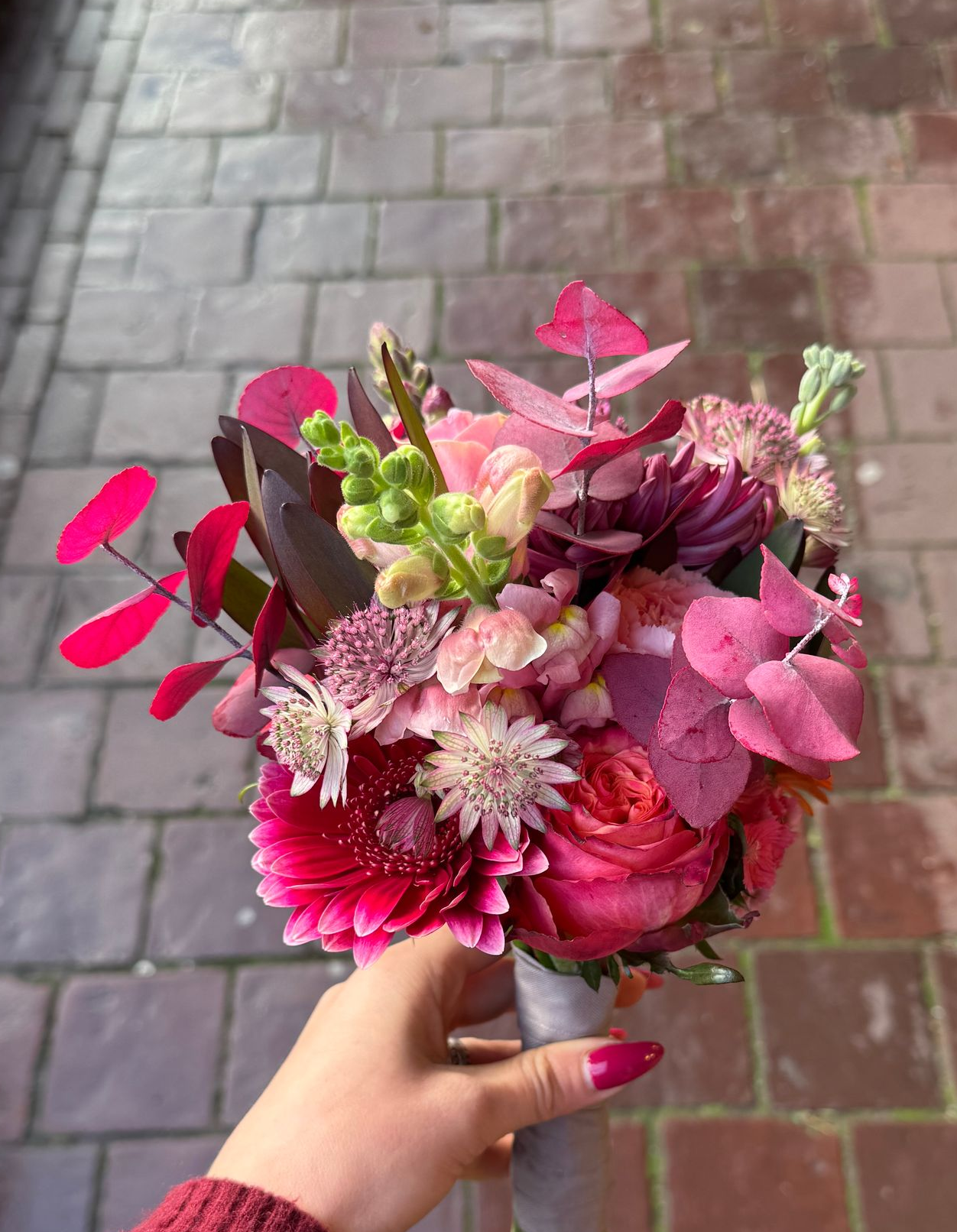 Een hand die een levendig boeket roze en paarse bloemen vasthoudt tegen een bakstenen achtergrond.