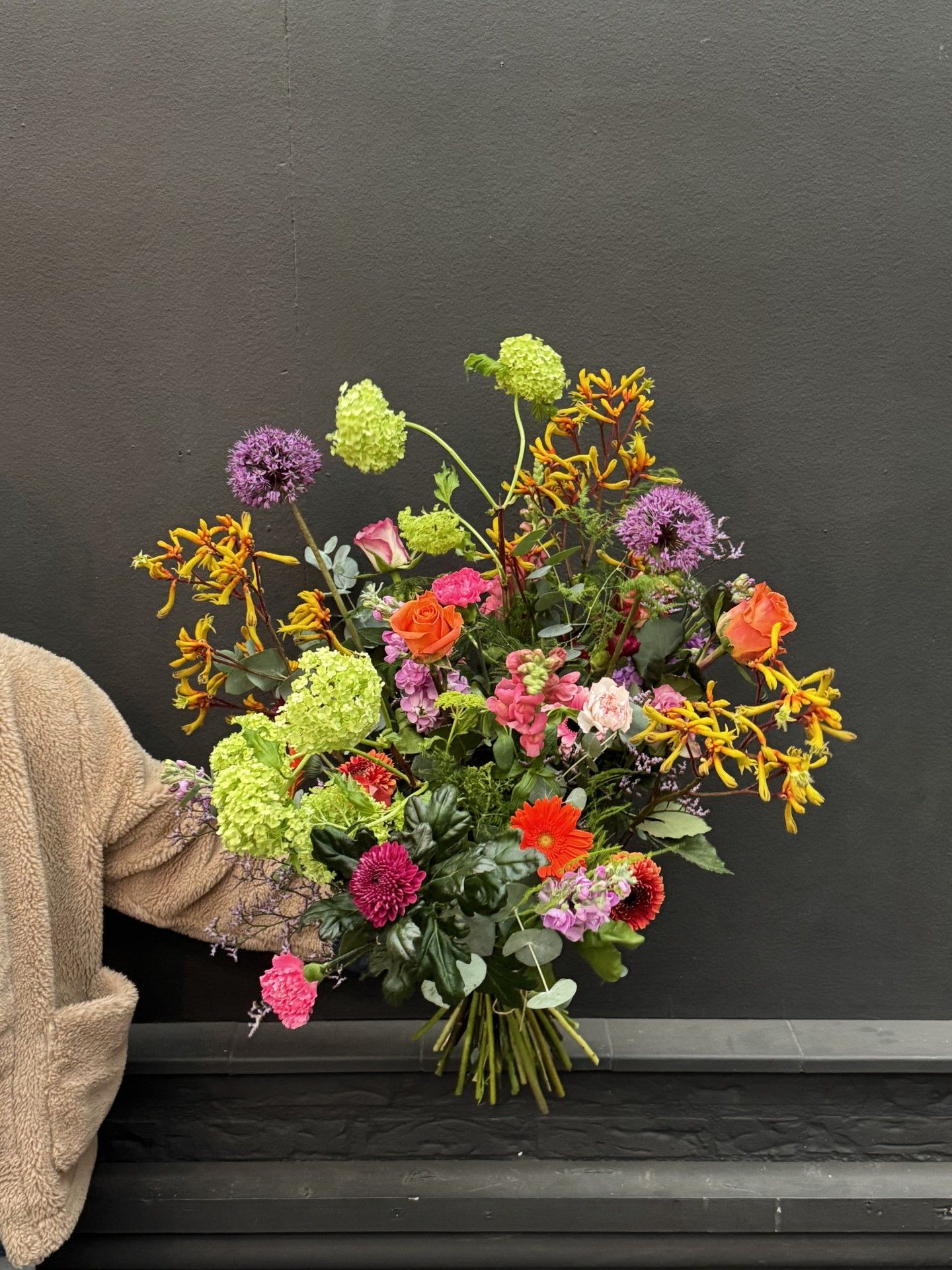 Een groot, levendig boeket met paarse, oranje, roze en limoengroene bloemen, tegen een donkergrijze muur.
