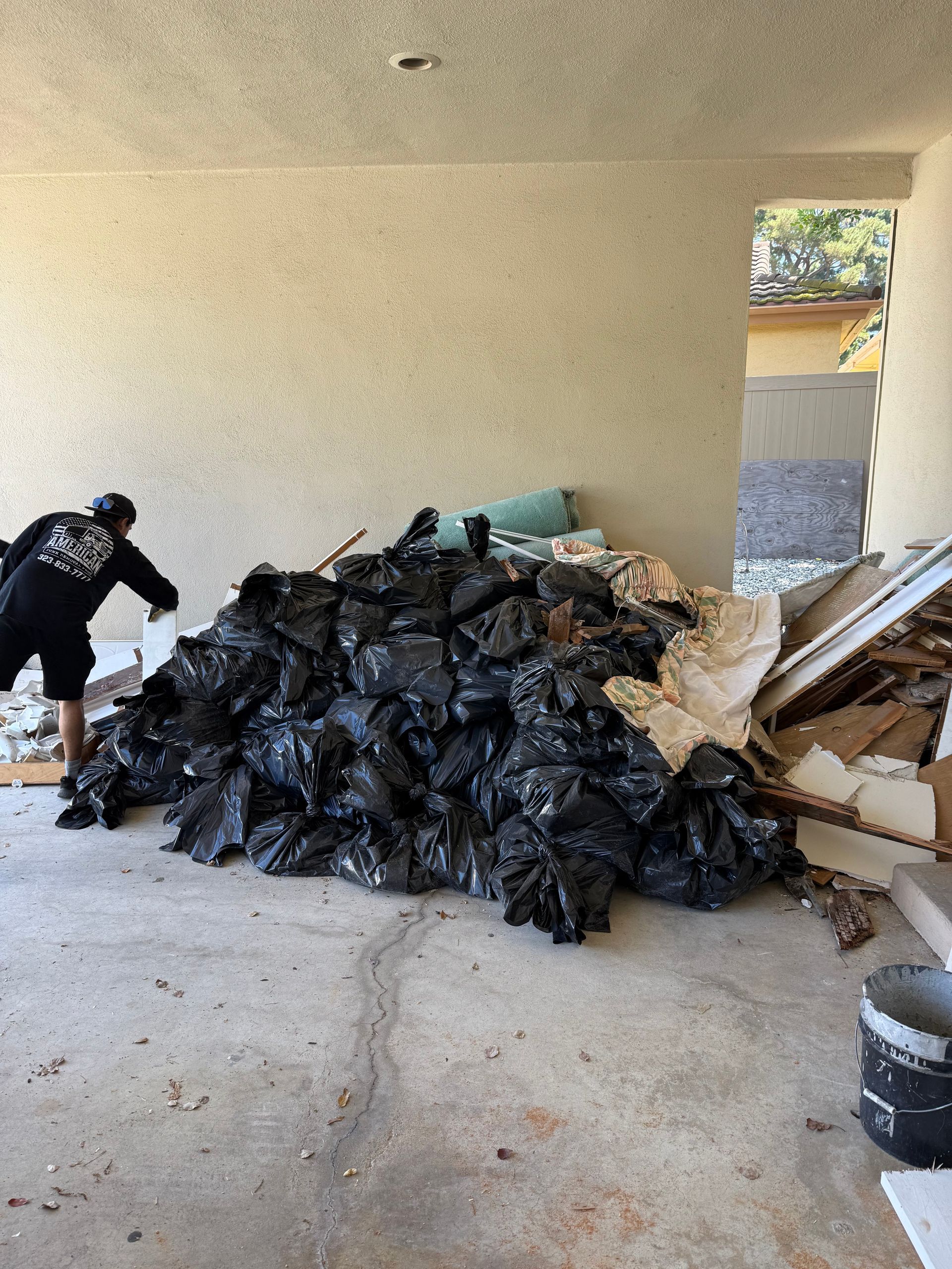 Person near large pile of black trash bags and debris in a concrete area, possibly a garage.