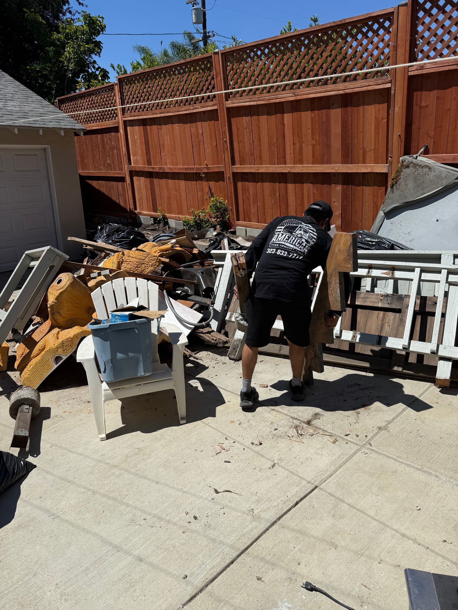 Person carrying wood near debris pile in a backyard. Wooden fence and shed visible in the background.