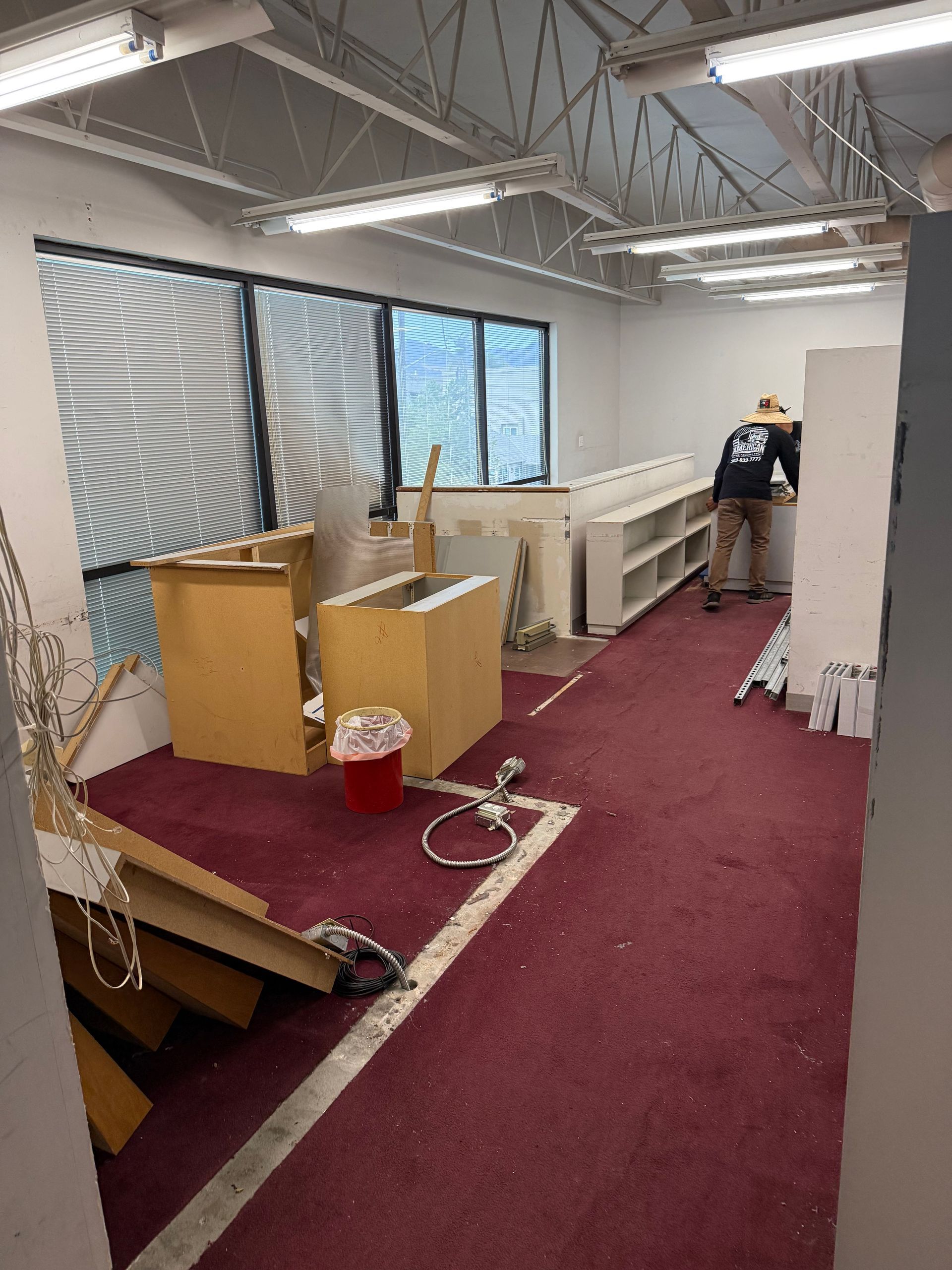 Interior view of a room with construction, a worker, boxes, cabinets, and burgundy carpet.