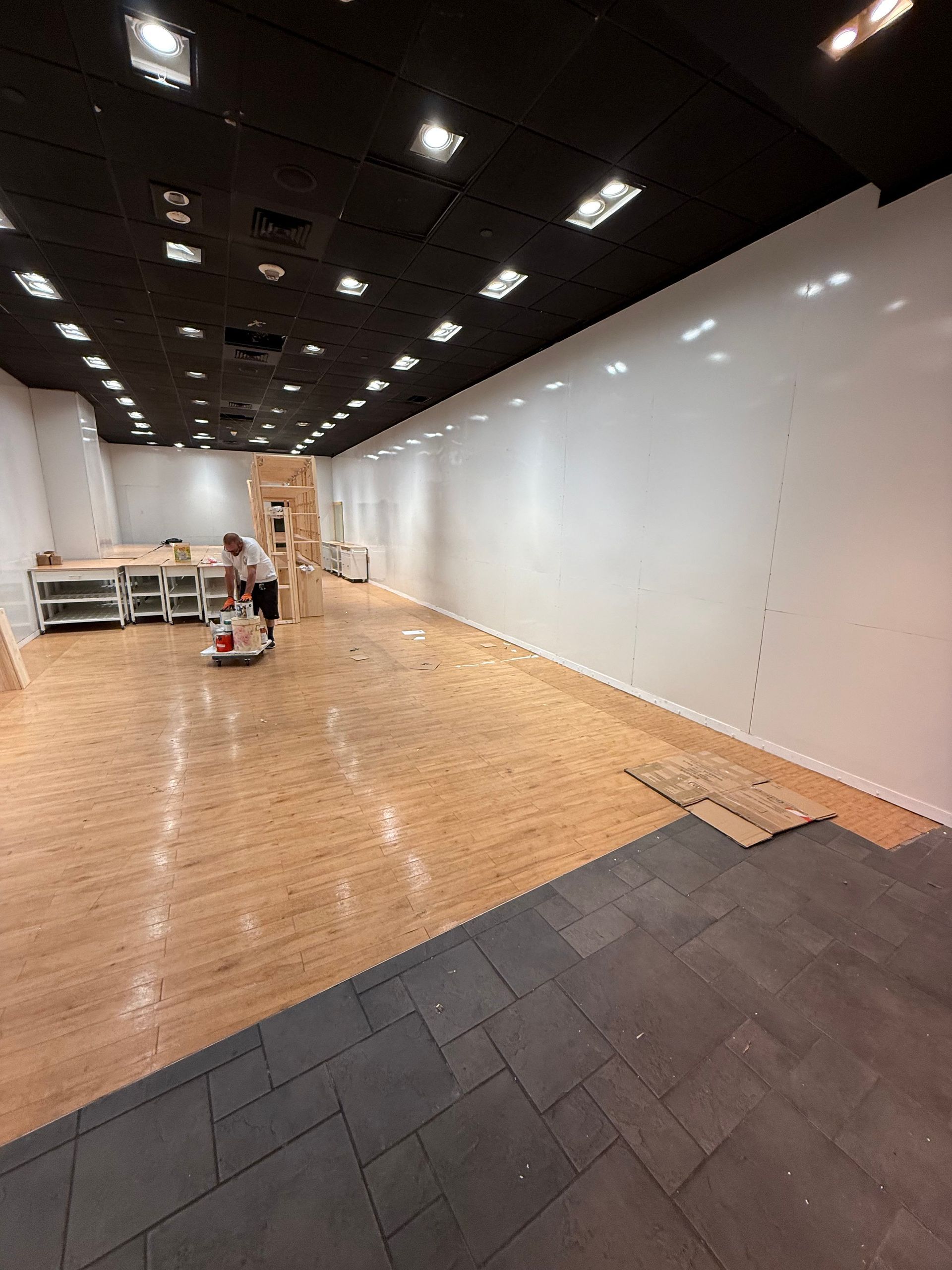 Interior of a space under construction; wood floor, white wall, black ceiling, person working.