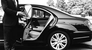 A person opening the rear door of a black luxury sedan in a driveway