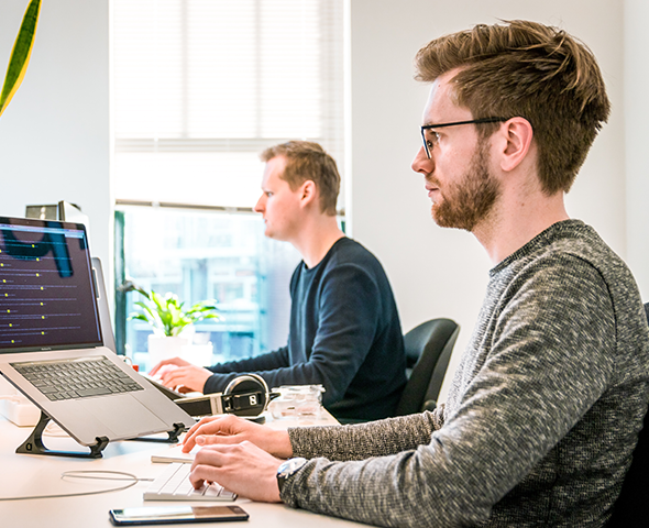 Two men are sitting at a desk working on laptops.