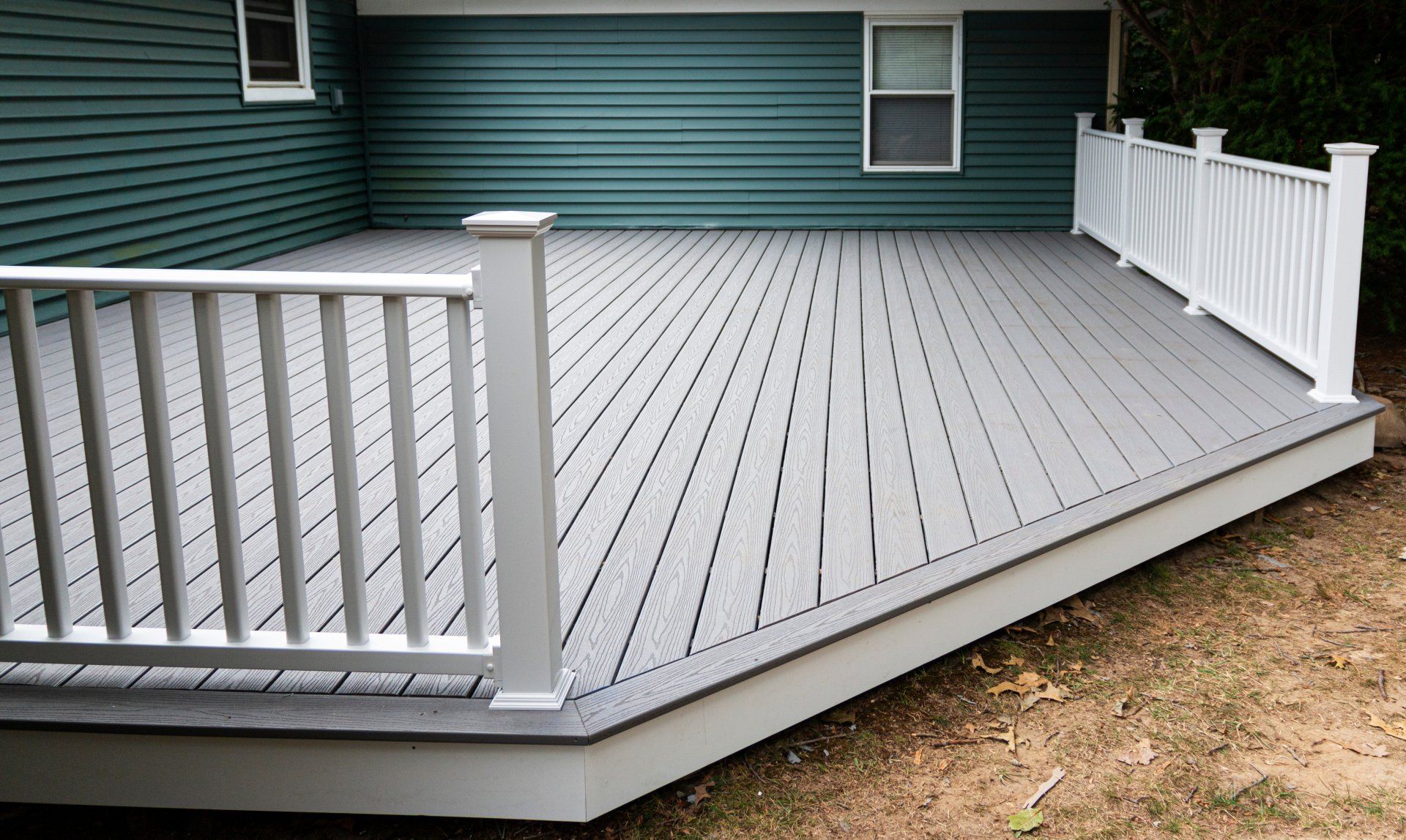 A gray deck with a white railing is in front of a green house.