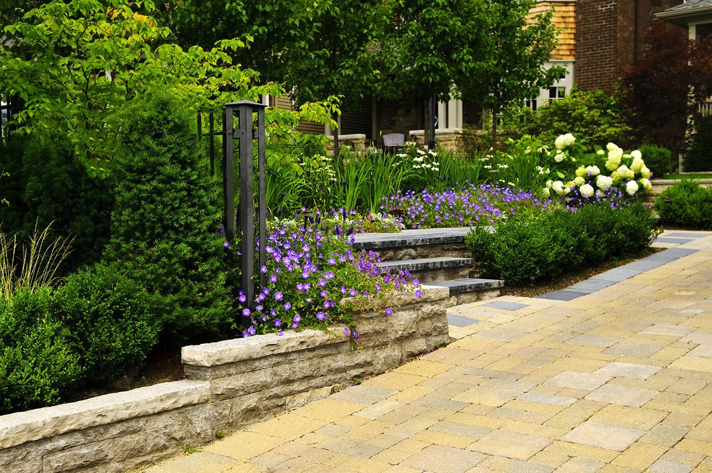 A brick walkway leading to a house surrounded by flowers and bushes