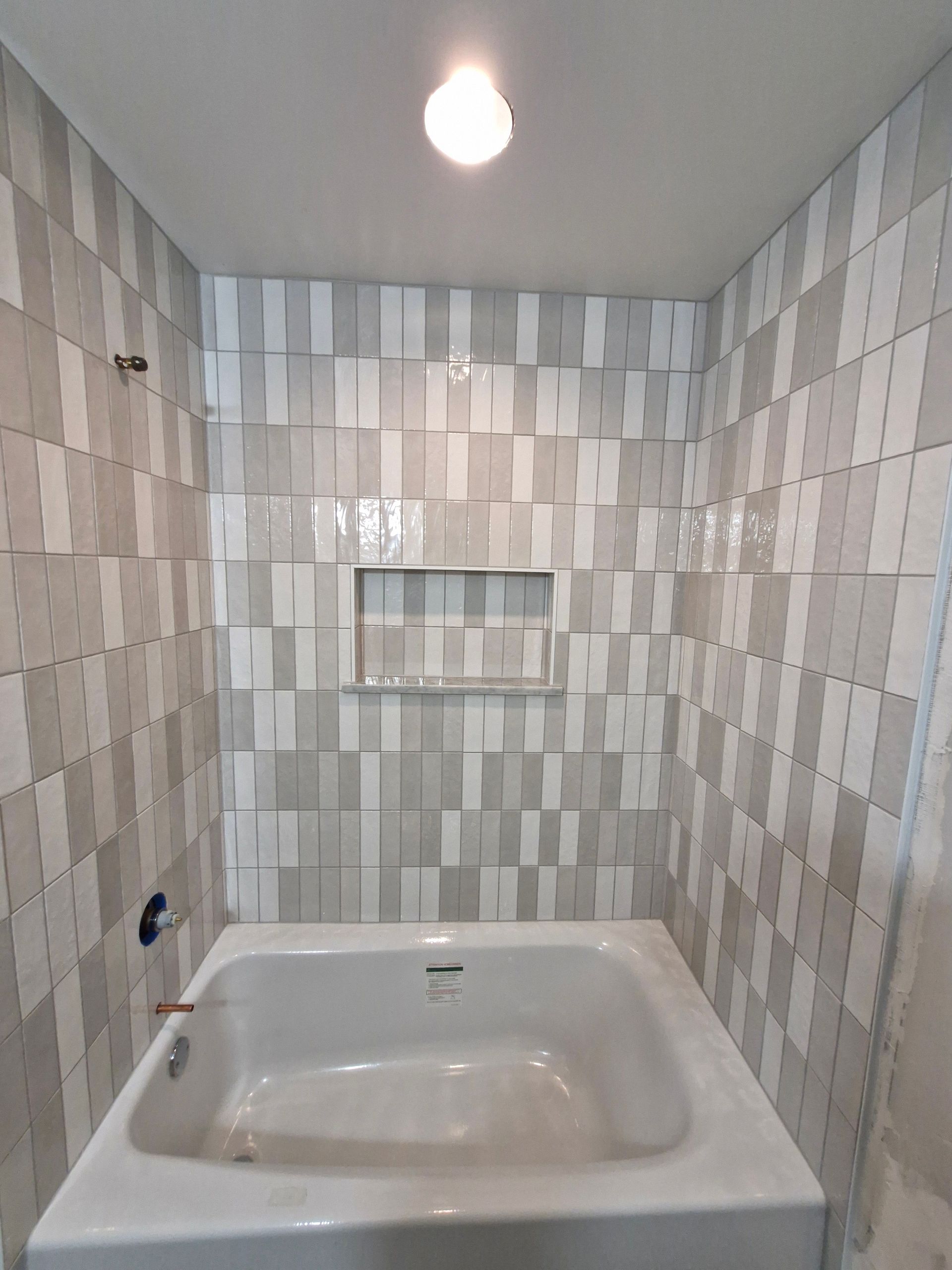 Bathroom with white bathtub, tiled walls in shades of gray and white, and built-in shelf.