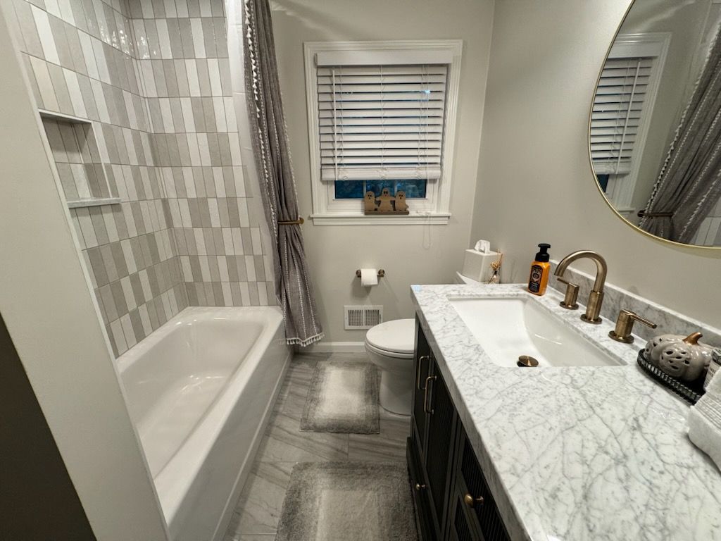 Bathroom with marble-patterned tile and countertop, dark vanity, bathtub, toilet, and a window with blinds.