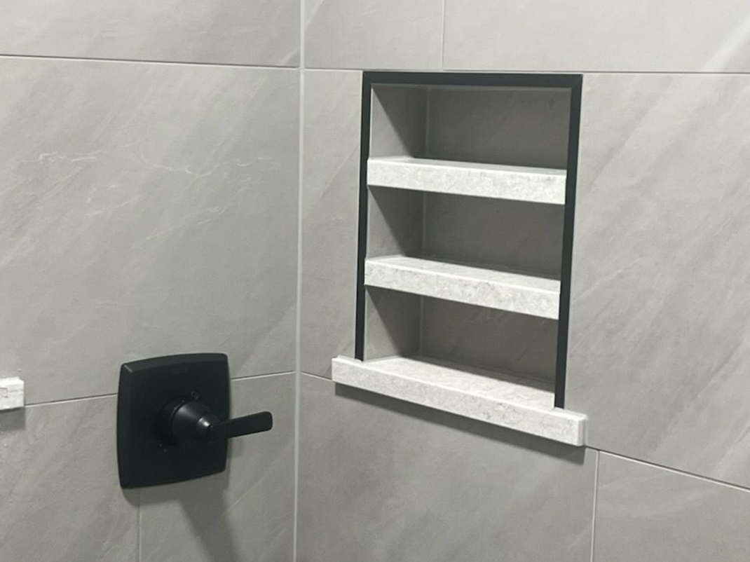 Bathroom with gray tiled walls, black faucet and a recessed shelf with three granite shelves.