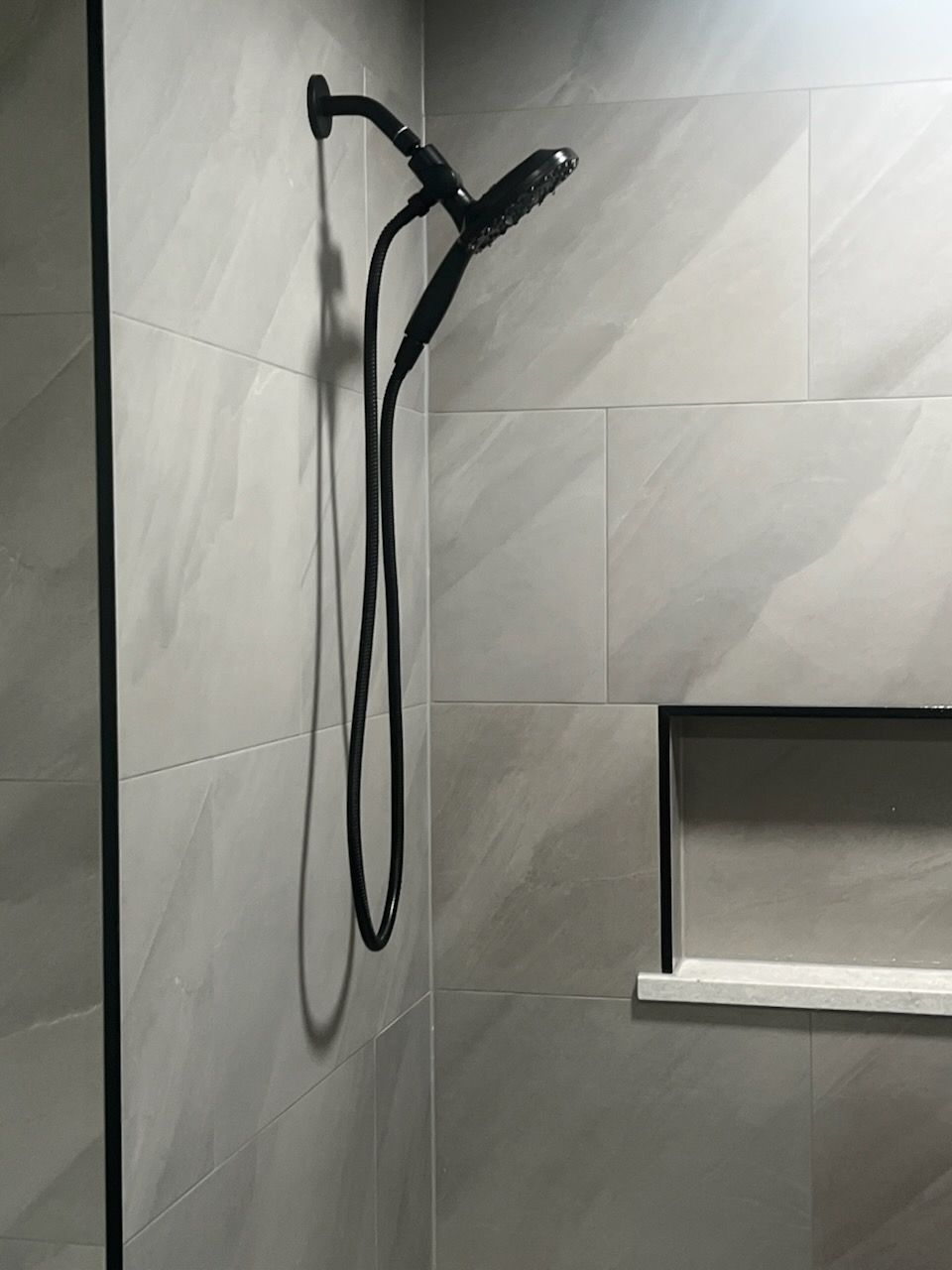 Black showerhead and hose against light gray tiled wall; a recessed shelf is visible.