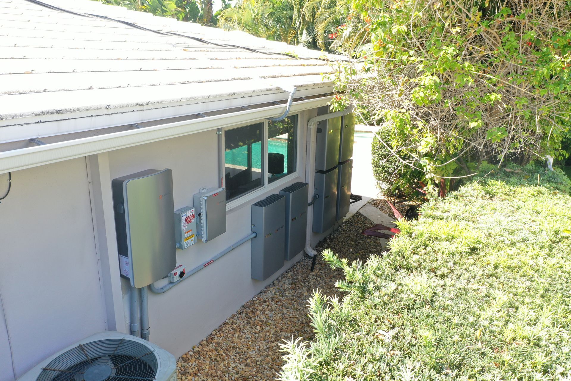 Side yard of a house with utility boxes, windows, and dense greenery beside a gravel strip