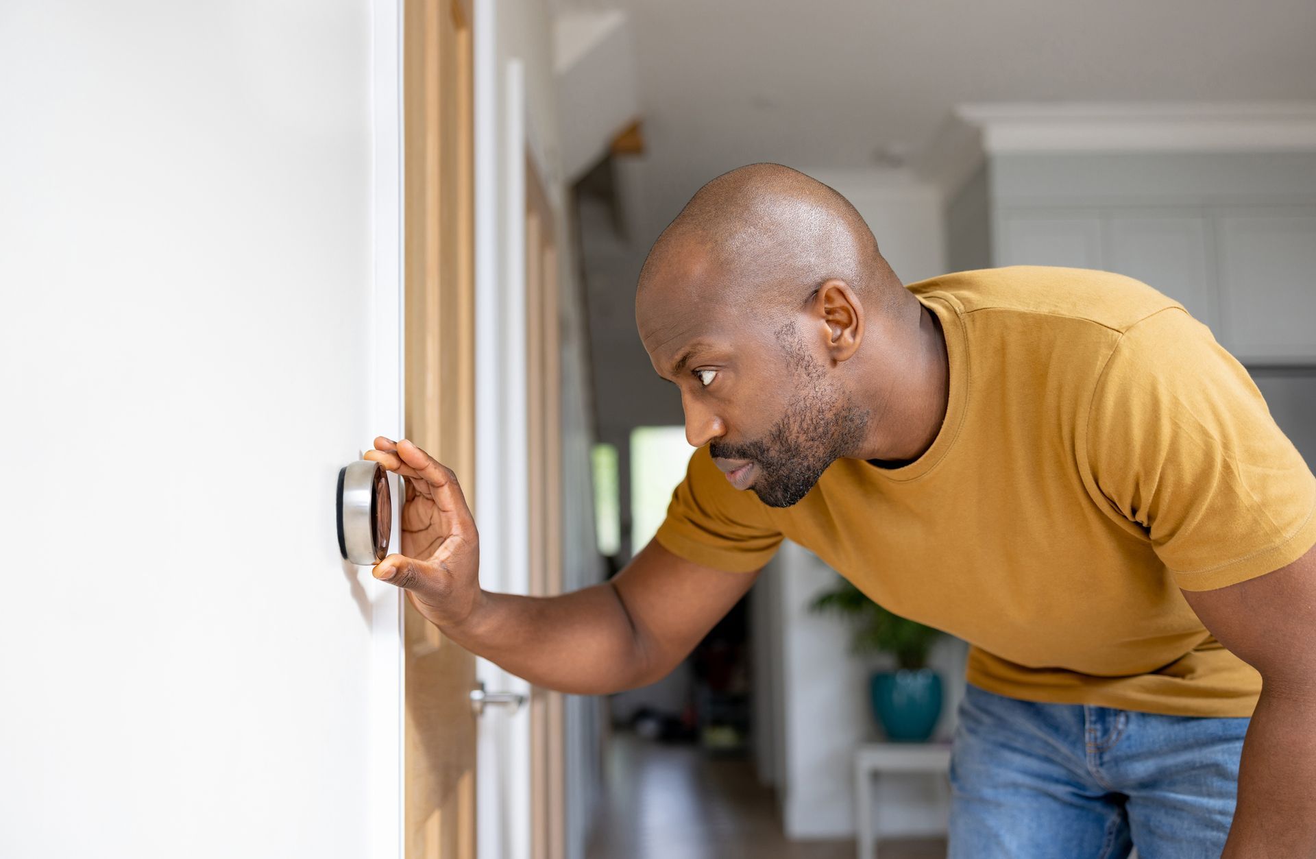Man adjusting a round, silver thermostat on a white wall. He wears a yellow shirt and jeans, indoors.