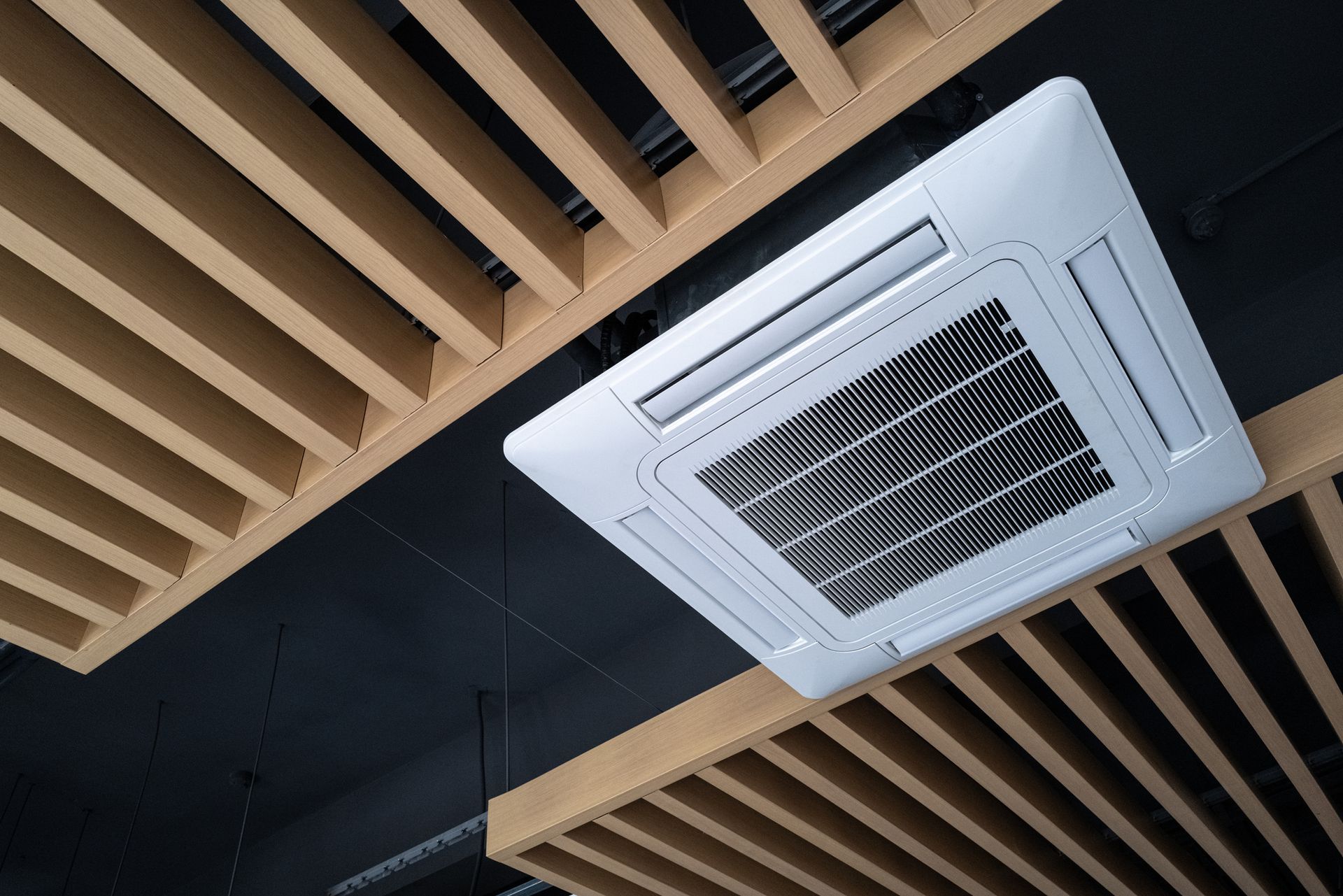 Ceiling-mounted air conditioning unit, white, with a dark ceiling and wooden beam design.