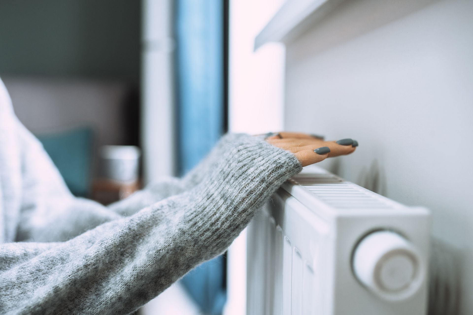 Person in gray sweater warms hands on a white radiator, indoors.