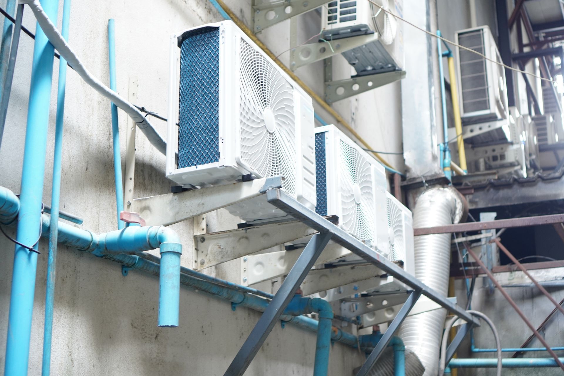 Air conditioning units mounted on an industrial wall with blue pipes, suggesting an outdoor or utility space.