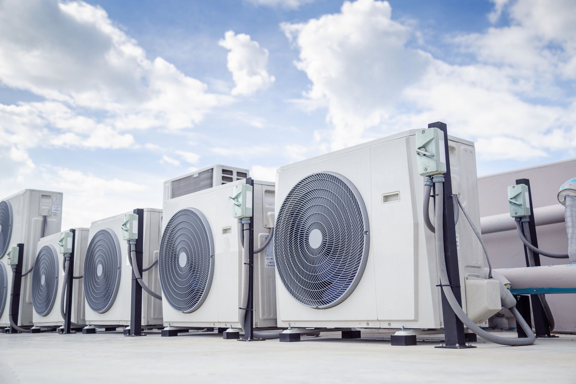 Air conditioning units lined up on a rooftop, against a cloudy blue sky.