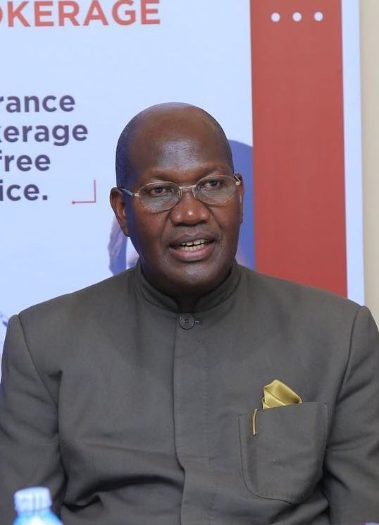 Man in suit, glasses, speaking. Brown jacket, gold pocket square, light background.