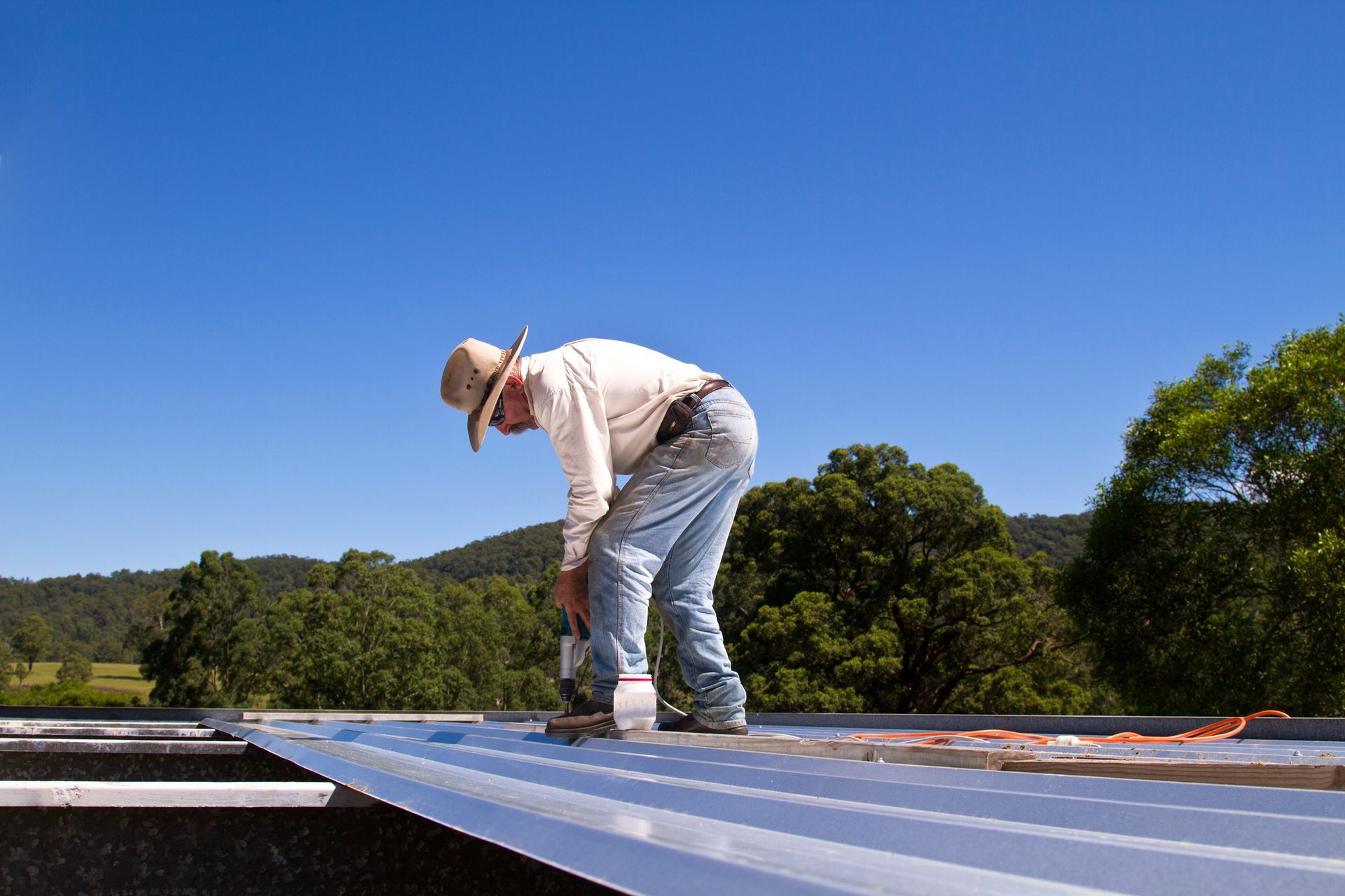 Roofer in jeans and hat, working on a metal roof with a drill under a blue sky.