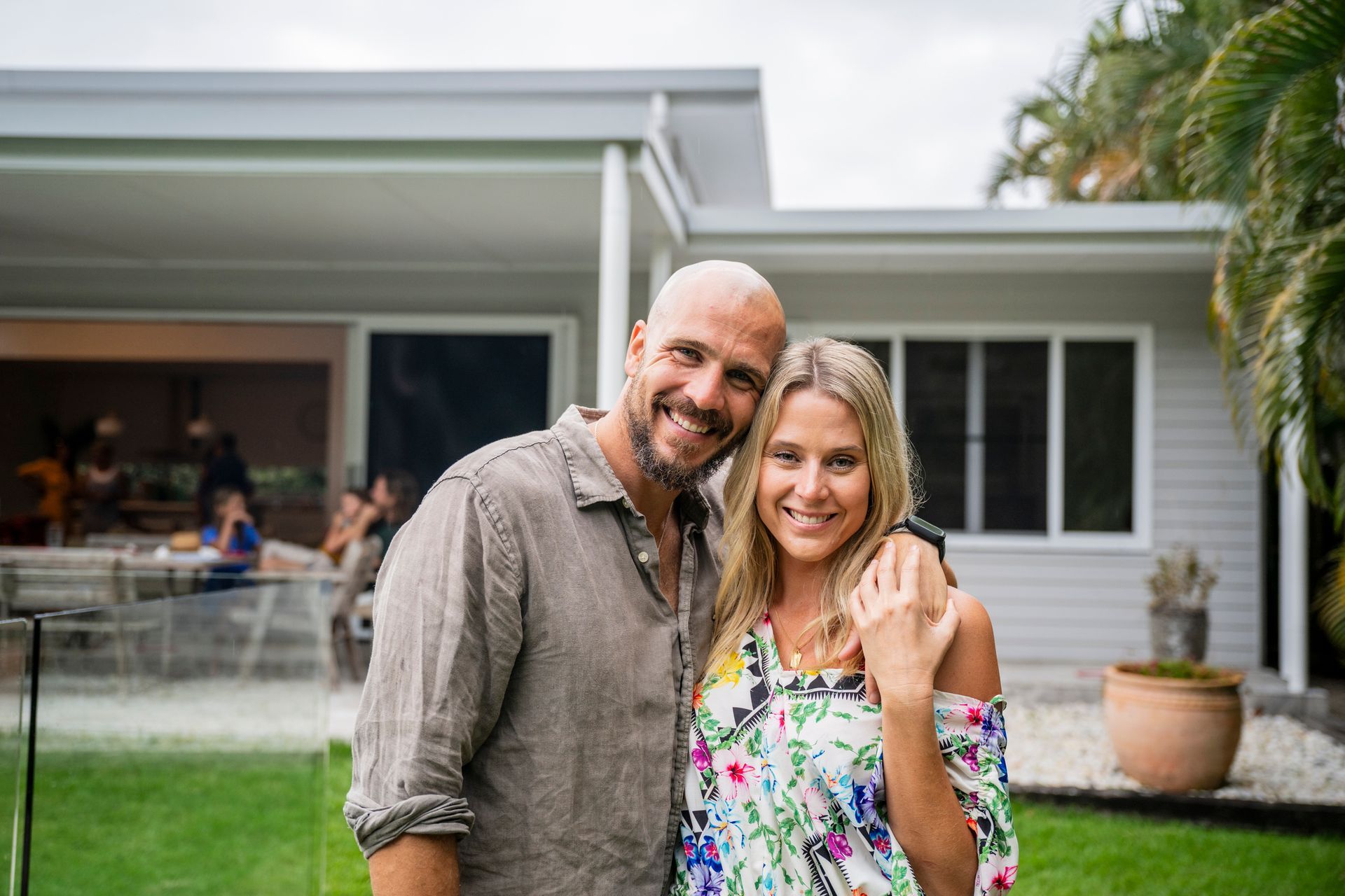 Smiling couple embracing in front of a house with a backyard, guests visible in the background.
