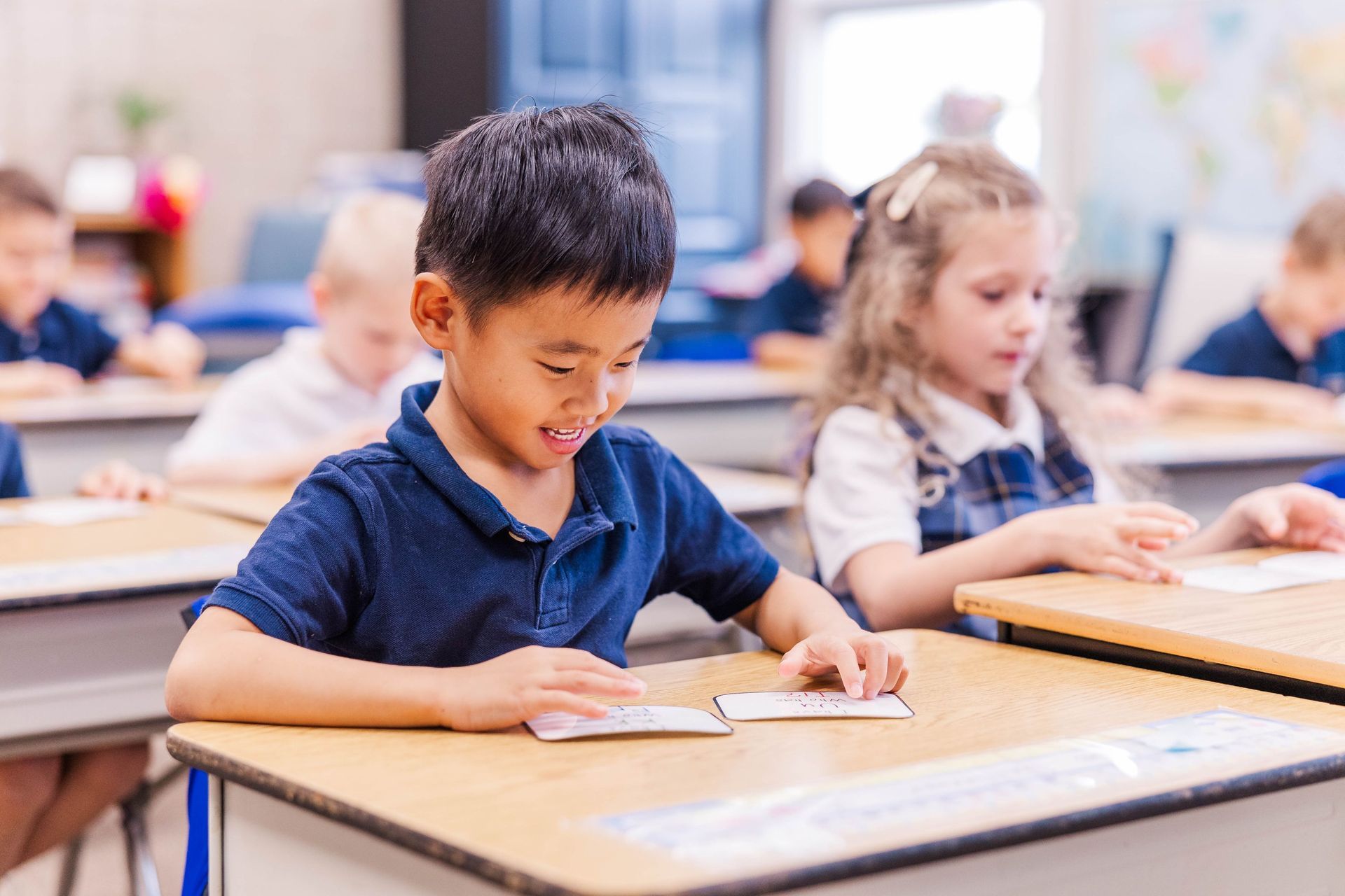 Boy in blue shirt smiles at cards on desk in classroom. Other students seated.