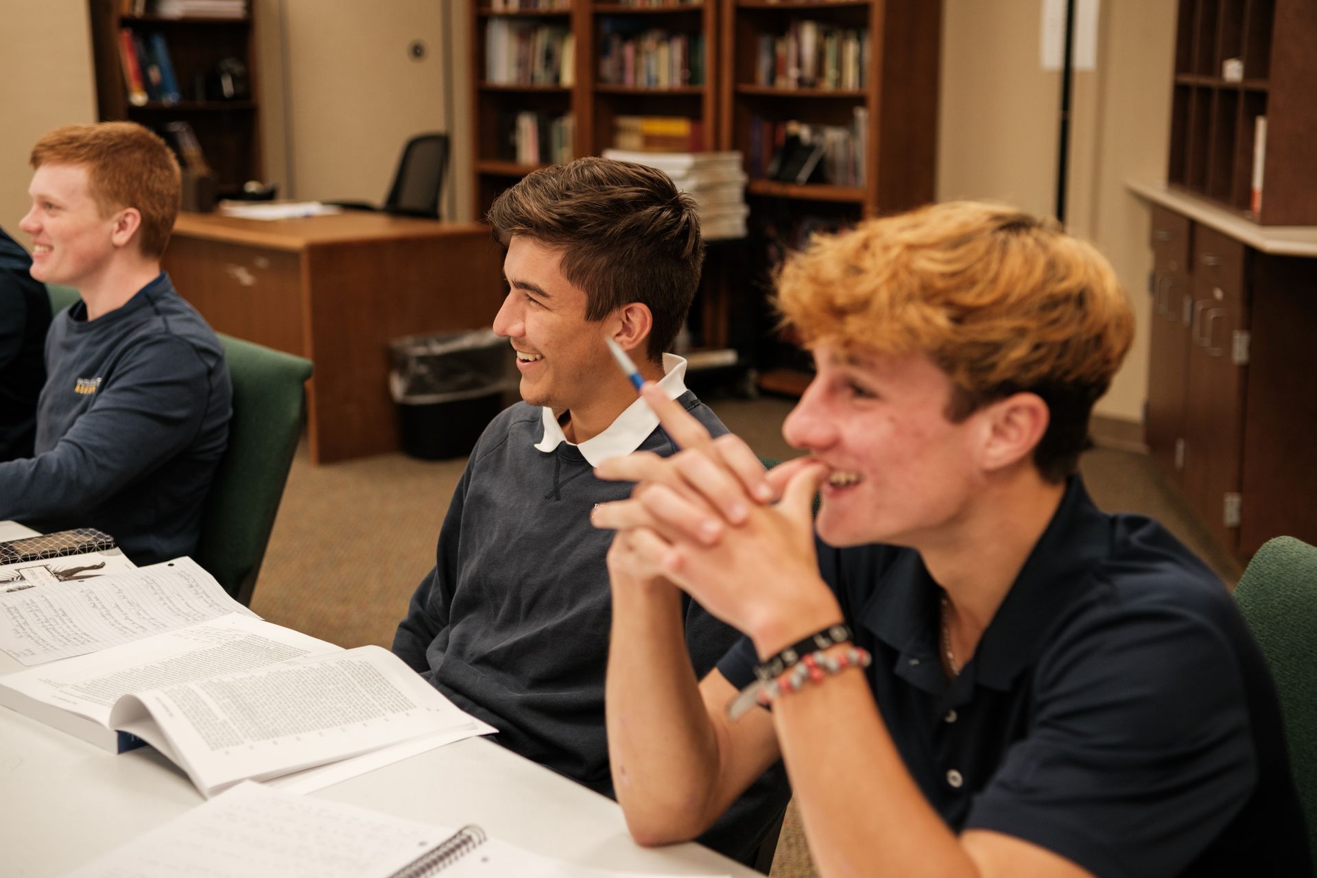 Three students seated at a table, studying. One smiles, hands clasped. Books and papers visible. Library setting.