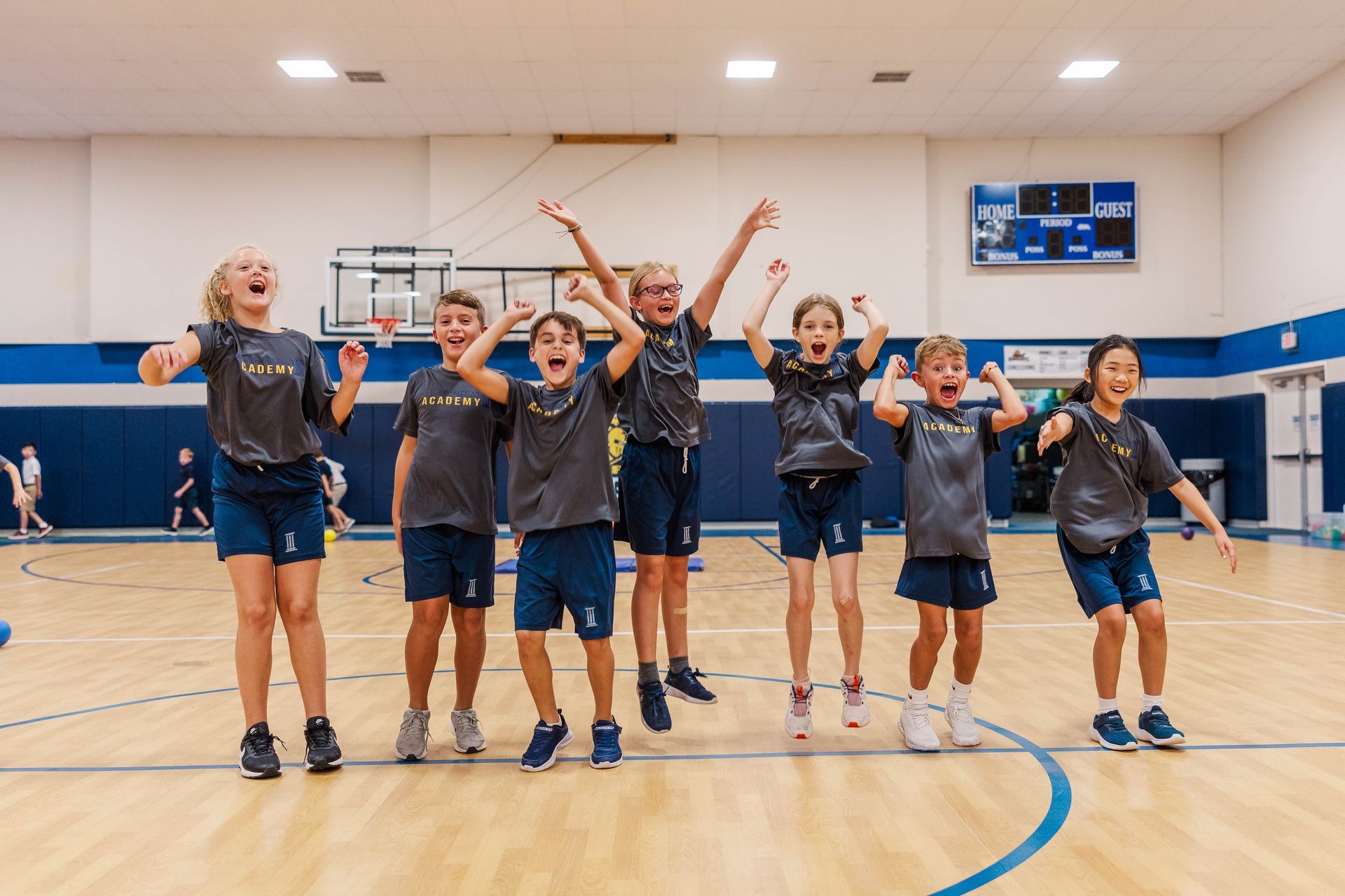 Children in gym jump and cheer, wearing gray shirts and blue shorts, hardwood floor, blue trim.