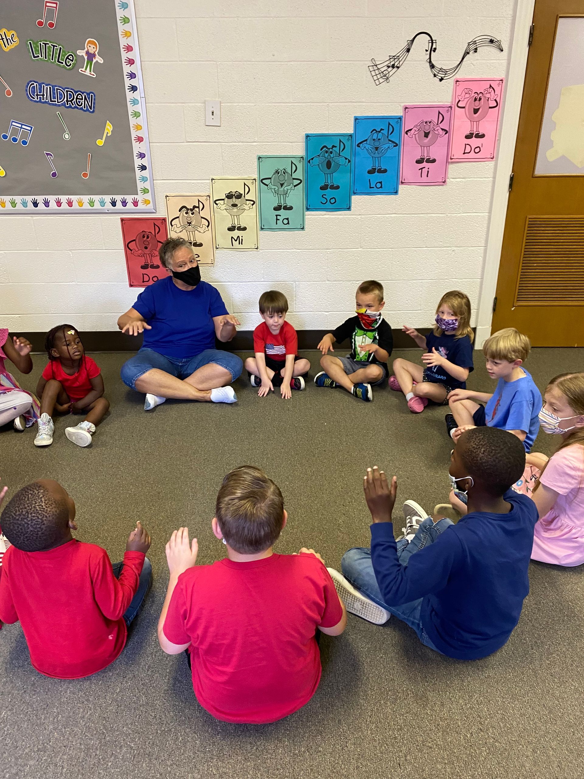 A group of children and an adult sit in a circle on the floor; they are in a classroom.
