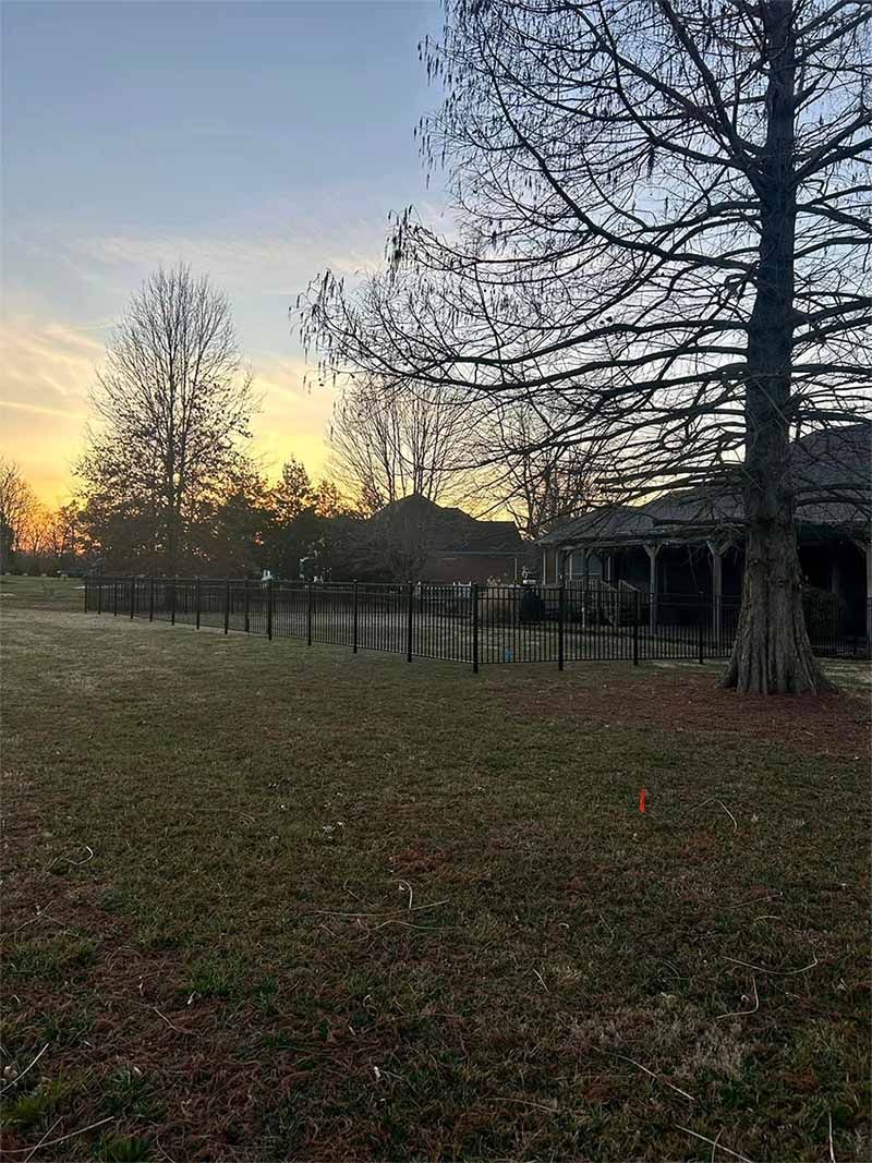 A fenced in yard with trees and a house in the background at sunset.