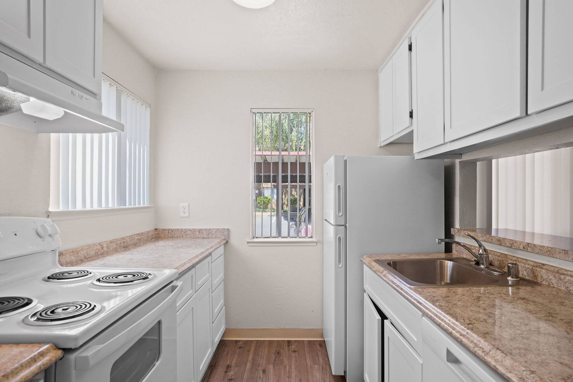 Bright white kitchen with granite counters, stove, sink, and a window with bars at the end.