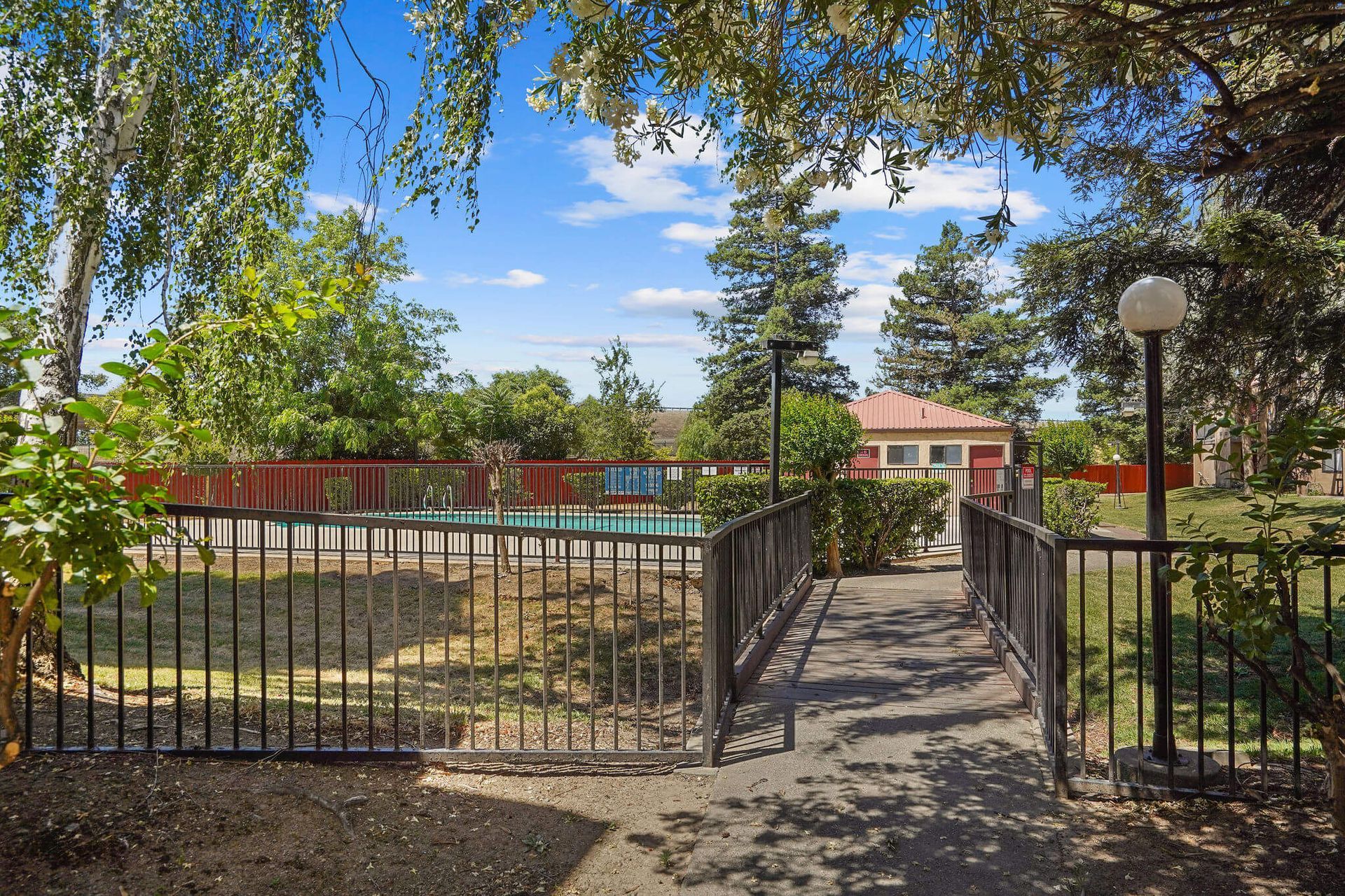Black iron gate leading to a sunny courtyard with a pool and red-roofed building under trees