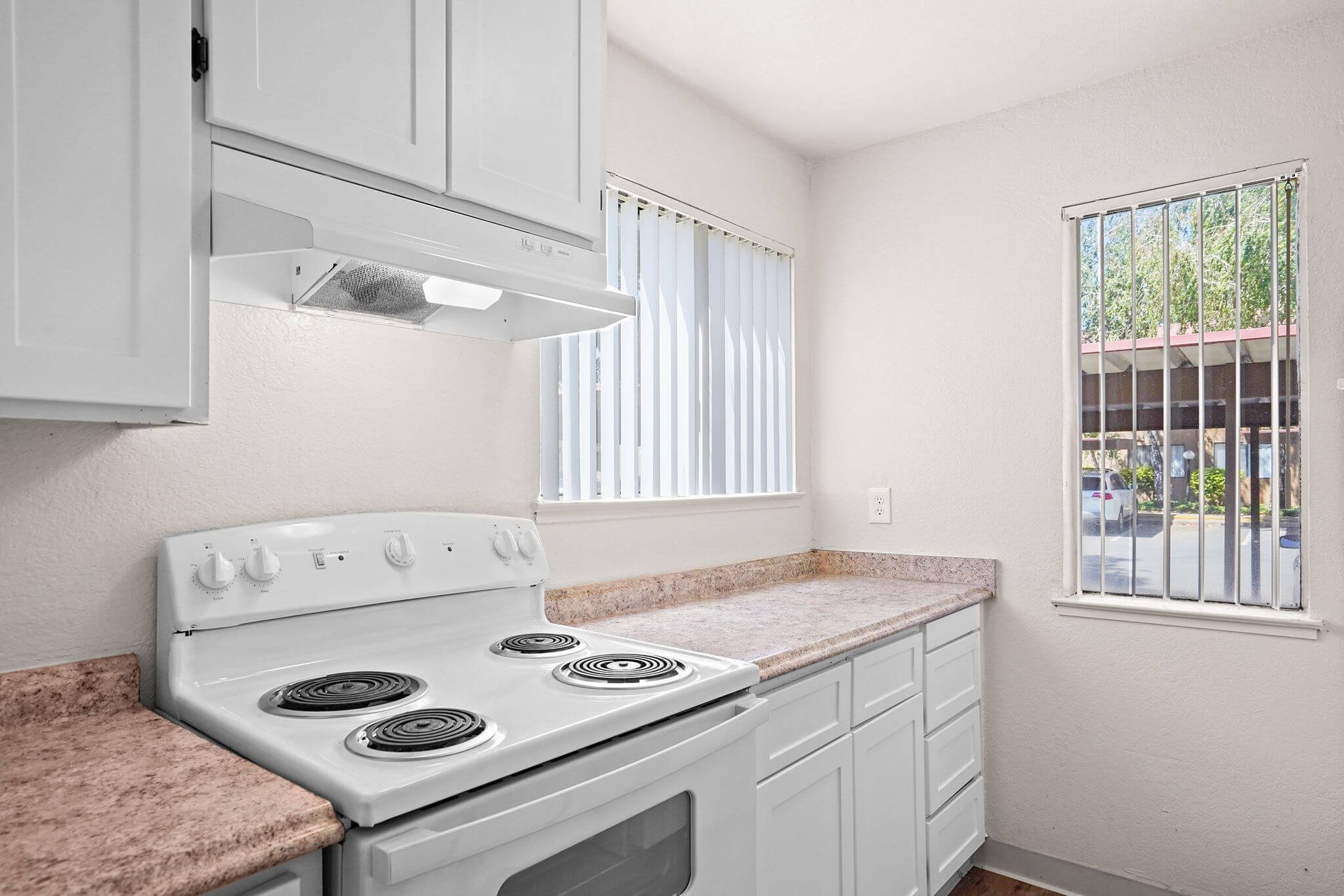 Bright white kitchen with electric stove, pink countertops, and two windows with vertical blinds