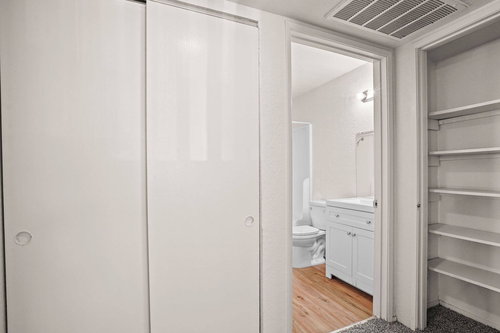 White hallway closet doors beside an open bathroom doorway with white cabinets and wood floor.