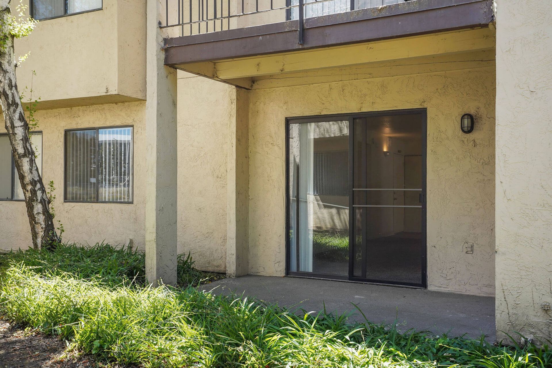 Apartment patio with sliding glass door, balcony above, and shrubs in front