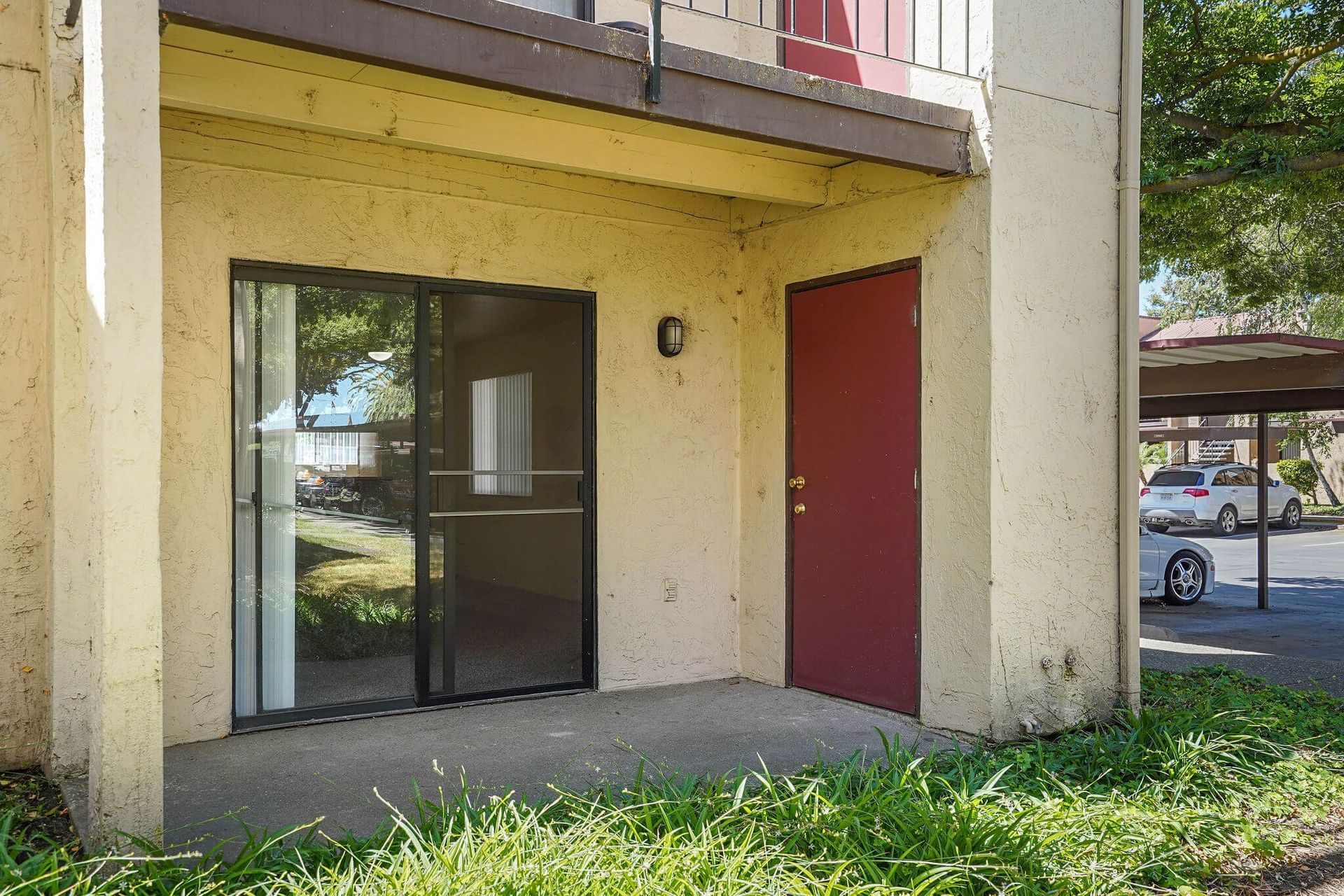 Apartment entrance with glass door, red side door, and small concrete patio under an overhang.