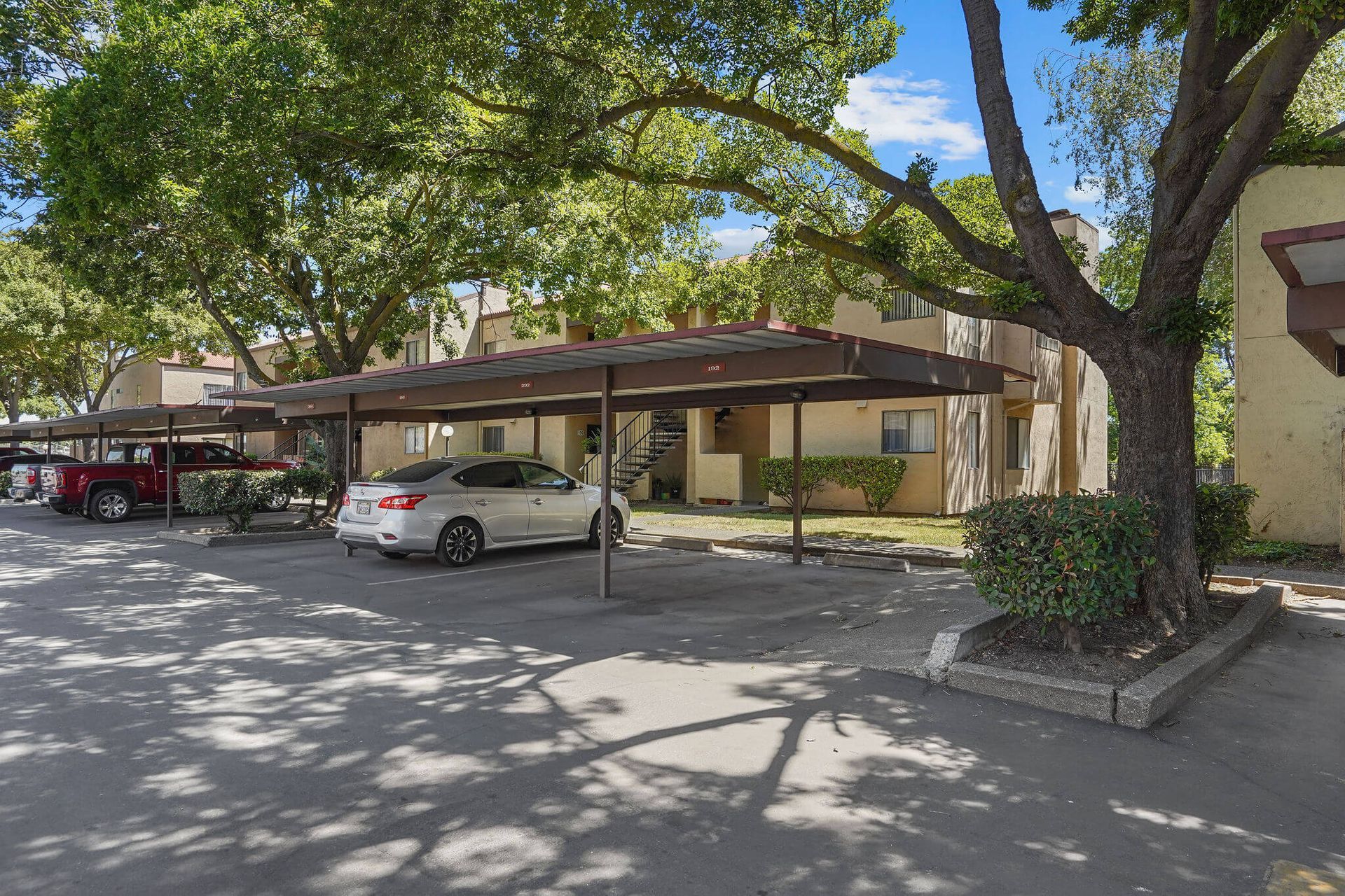 Apartment parking lot with covered carports, parked cars, and large trees shading the lot