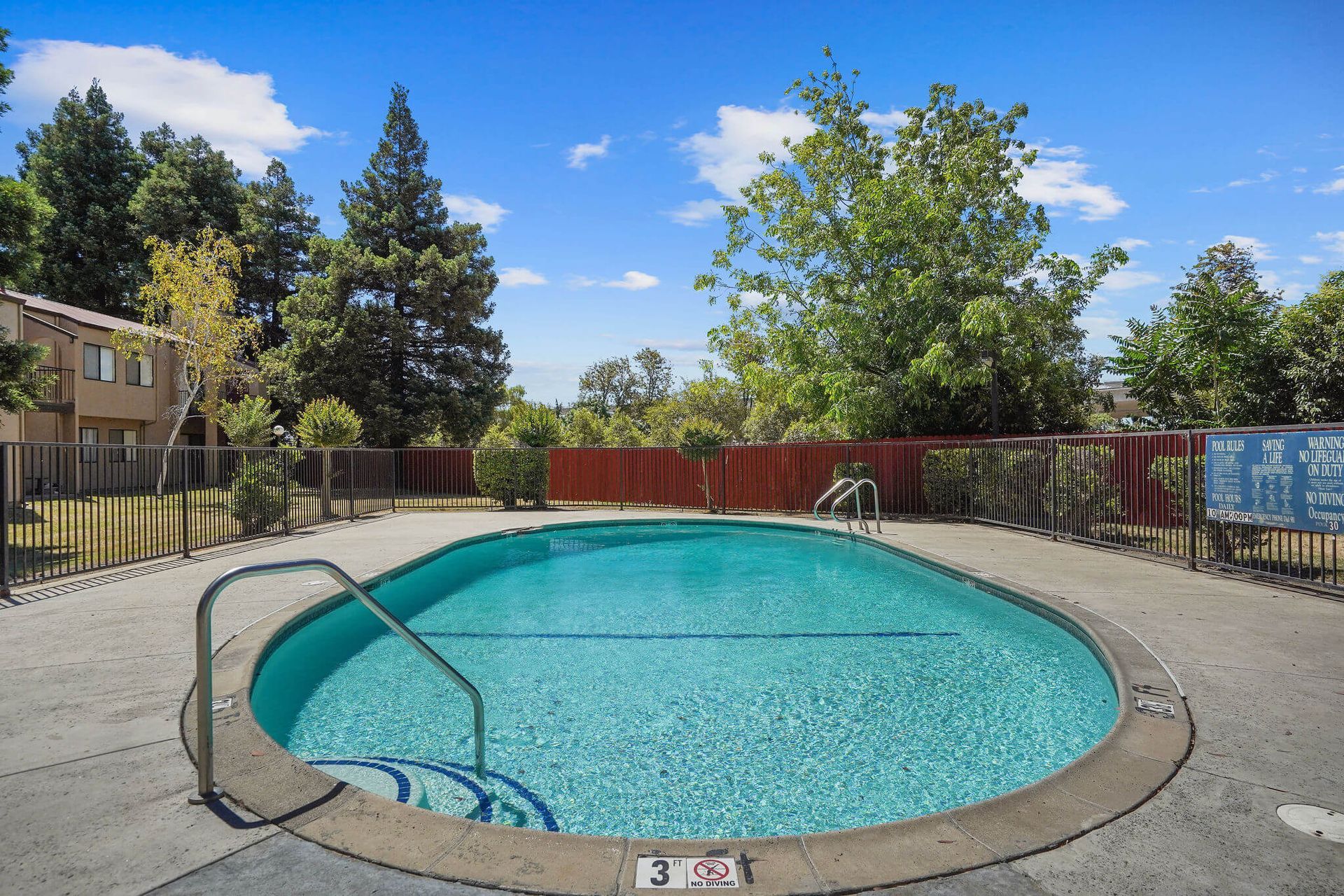 Outdoor apartment pool with blue water, concrete deck, metal railing, and trees under a bright sky