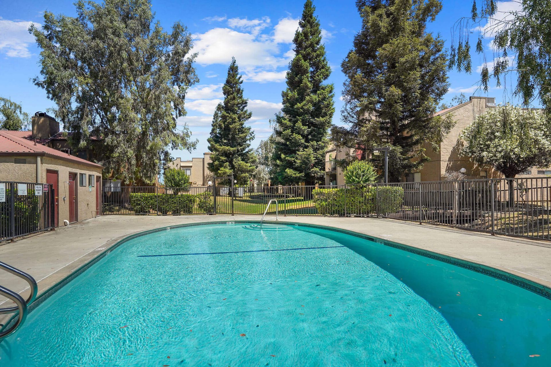 Outdoor swimming pool with clear blue water, surrounded by trees, apartments, and a fence under a sunny sky