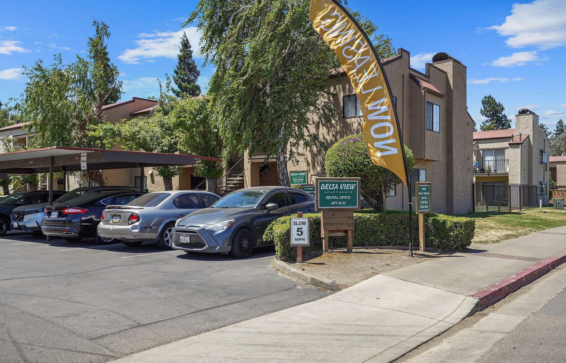 Apartment complex parking lot with cars, trees, and a tall sign by the sidewalk