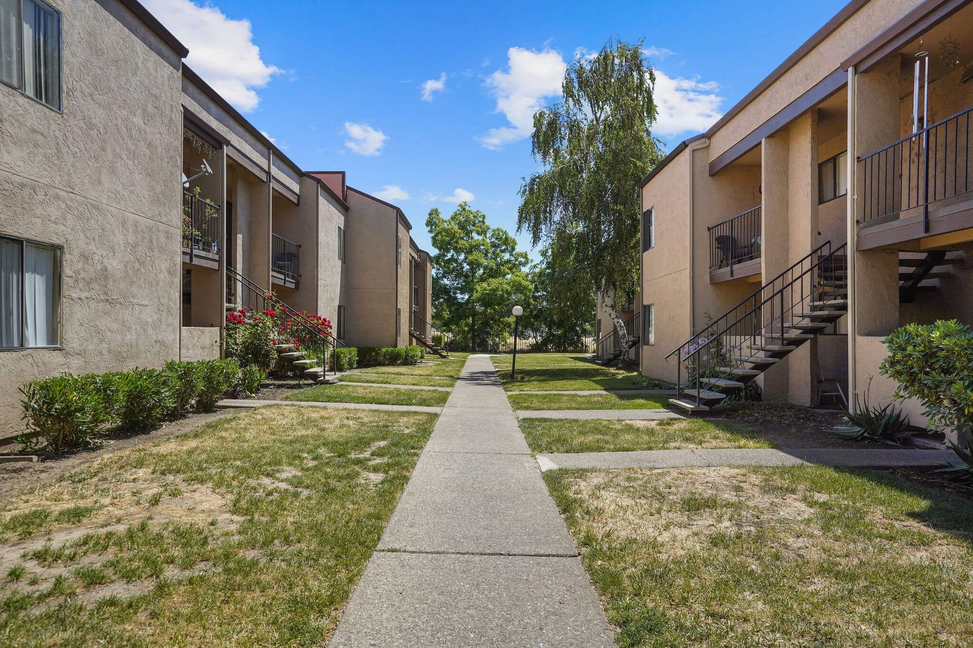 Apartment courtyard with central walkway, grass, and beige buildings under a bright blue sky