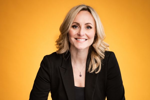 A woman in a black jacket is smiling in front of a yellow background