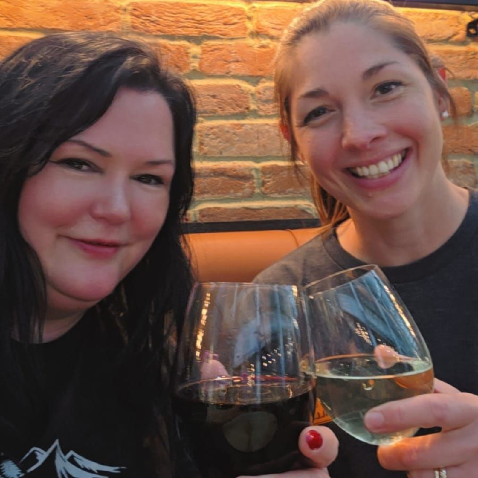 Two smiling women toasting with wine glasses in front of a brick wall.