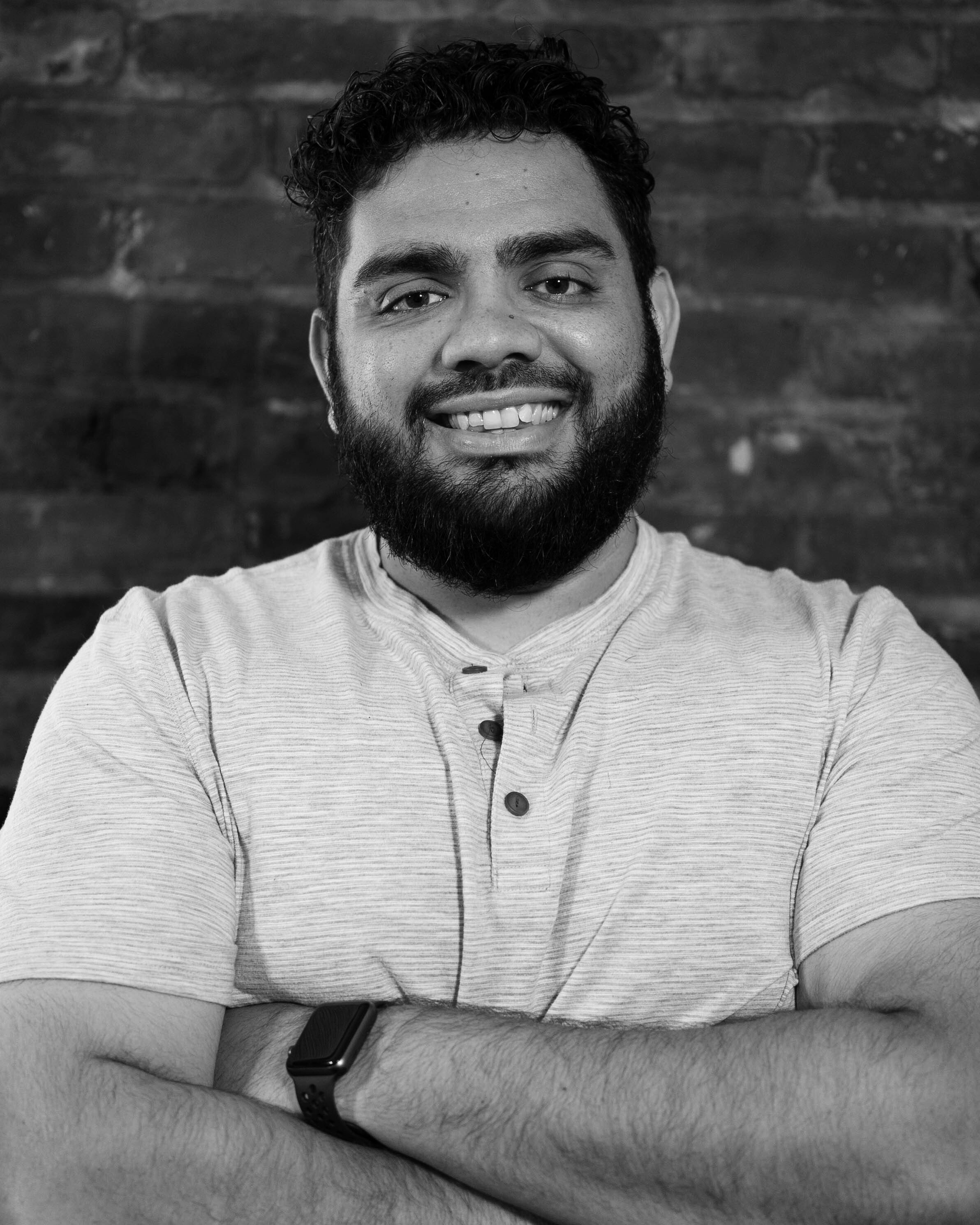A man with a beard is smiling in a black and white photo with his arms crossed.